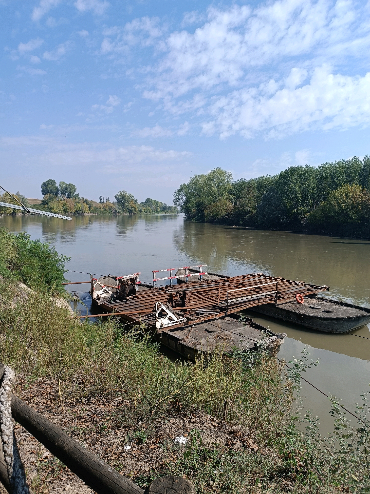 The image shows a river scene with a rusty barge tied to the bank in the foreground. The barge appears to be composed of multiple pontoons and has a wooden deck with metal railings and winches. There is a life preserver attached to the barge. The river is calm and reflects the sky, which is partly cloudy and blue. The banks of the river are lined with trees and vegetation. In the distance, there is a ramp-like structure extending from the left bank into the river. The scene appears to be in Marcaria, Italy, likely near a ferry crossing point. There are no people visible in the image.