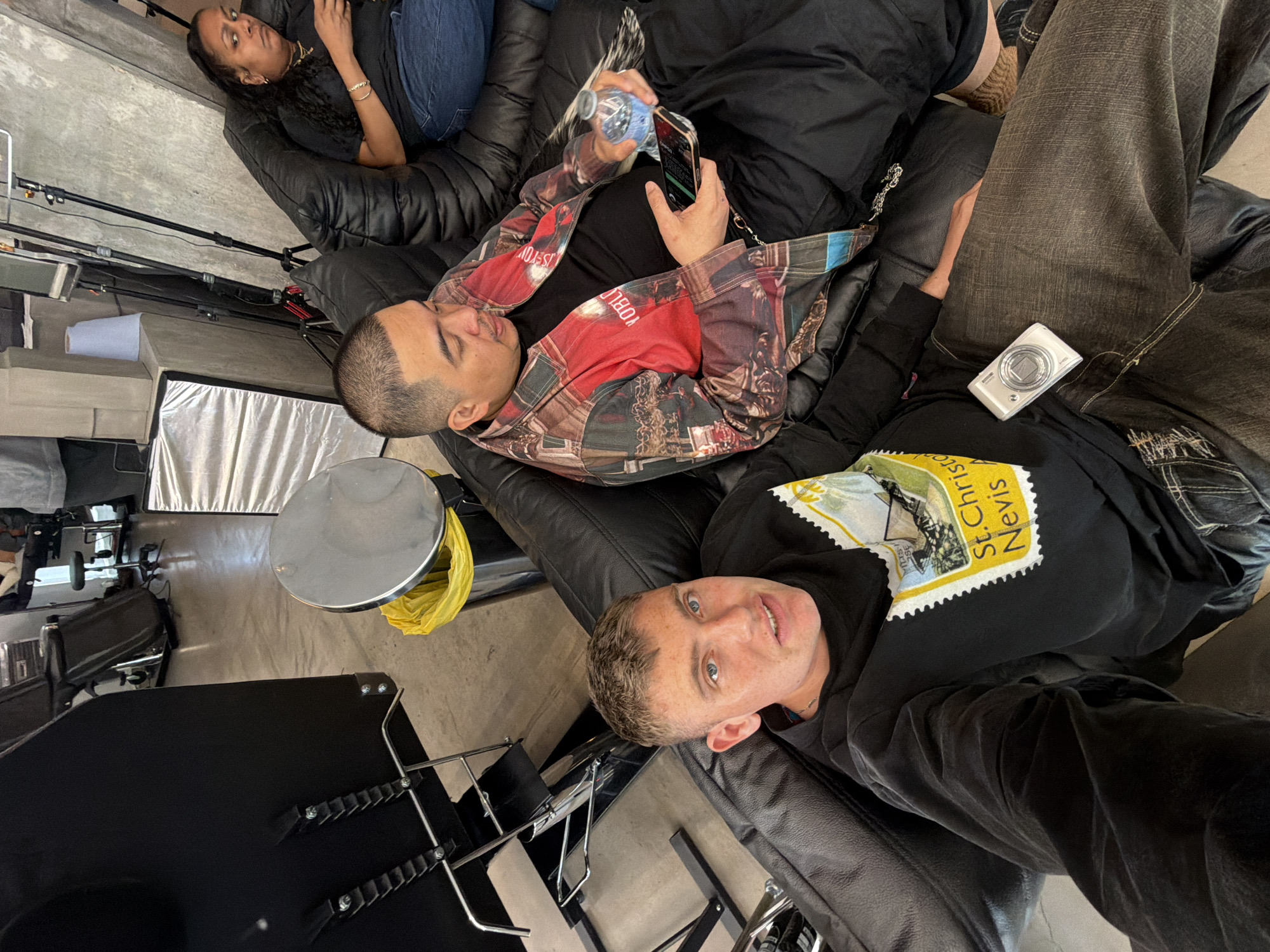 This rotated image captures three individuals relaxing on black leather seating in an indoor setting, likely a studio or lounge area in London, United Kingdom.
In the background, a woman with long, dark, braided hair, dressed in a black top and blue jeans, reclines on the couch. In the mid-ground, a man with short dark hair wears a striking red and black patterned long-sleeved shirt and black pants. He is engaged with his smartphone held in one hand and a water bottle in the other. Partial text on his shirt includes fragments like "IS YOU" and "YOBLE."
Closer to the viewer, a man with short light-colored hair and striking blue eyes lies on the seating, looking directly at the camera with a neutral expression. He is wearing a black long-sleeved shirt adorned with a prominent yellow and white graphic designed like a postage stamp, clearly displaying the text "St. Christoph Nevis." A small, white digital camera rests on his thigh.
The environment suggests a functional space, possibly a photography studio, with visible equipment such as light stands and a softbox. A round silver table or stool and a yellow plastic bag are also present in the scene. The floor is a light-colored concrete or industrial surface. The overall impression is one of a casual break or downtime in a creative or professional context.