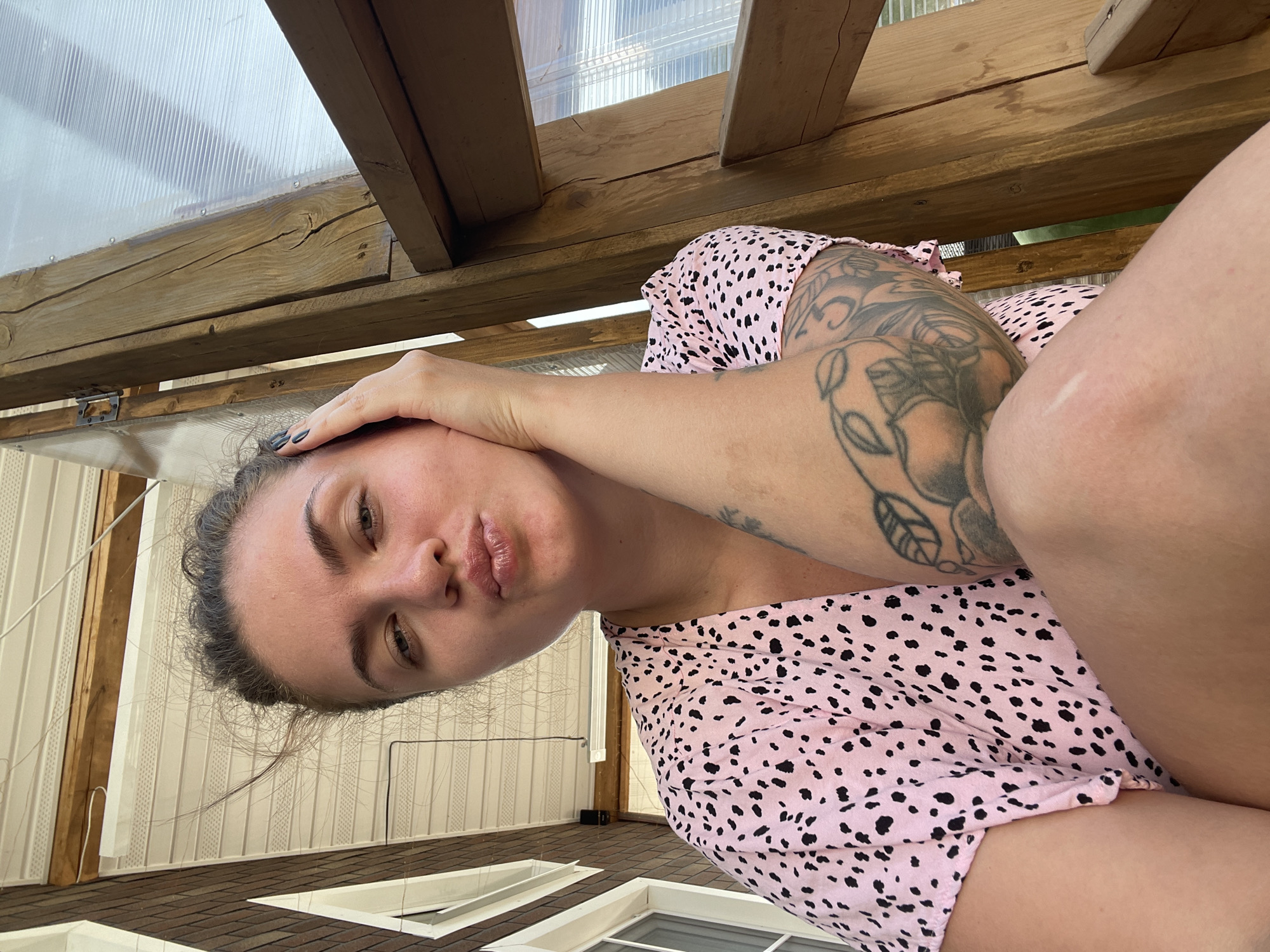 A woman is reclining in a partially constructed or unfinished greenhouse or sunroom. She wears a pink top with black spots and has a tattoo on her left arm. Her left hand rests on her forehead. The structure surrounding her consists of wooden beams and a translucent roof. The walls appear to be constructed of white siding with dark trim.