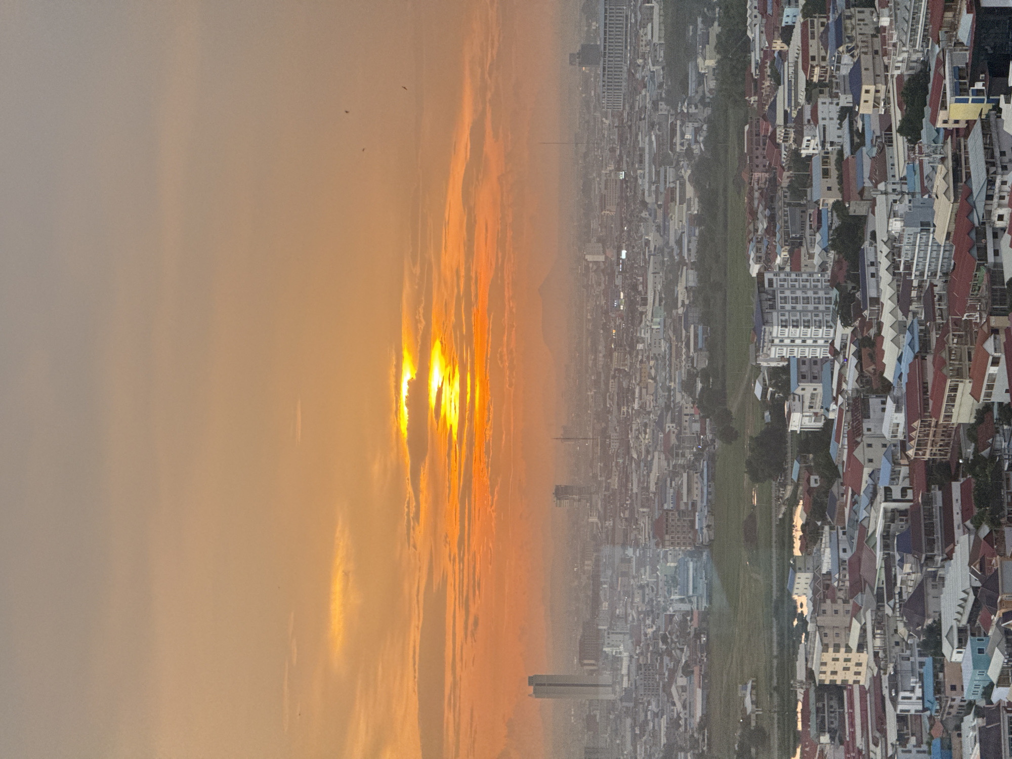 The image captures a wide aerial view of a densely populated urban landscape in Khan Mean Chey, Cambodia, during what appears to be either sunrise or sunset. The sky is dominated by warm hues of orange and yellow, with clouds creating a textured backdrop. The sun is partially visible, casting a soft glow over the scene. Below the sky, a sprawling city unfolds, characterized by a multitude of buildings of varying heights and architectural styles. Many buildings feature red or blue roofs, interspersed with white and beige facades. There are several taller structures, including at least two prominent skyscrapers, scattered throughout the cityscape. A large green field, possibly a park or undeveloped land, is visible in the middle ground, with a body of water reflecting the sky's colors. The distant horizon is hazy, with faint outlines of mountains or larger structures. The overall impression is one of a bustling city at the beginning or end of a day, with a mix of modern development and more traditional residential areas.