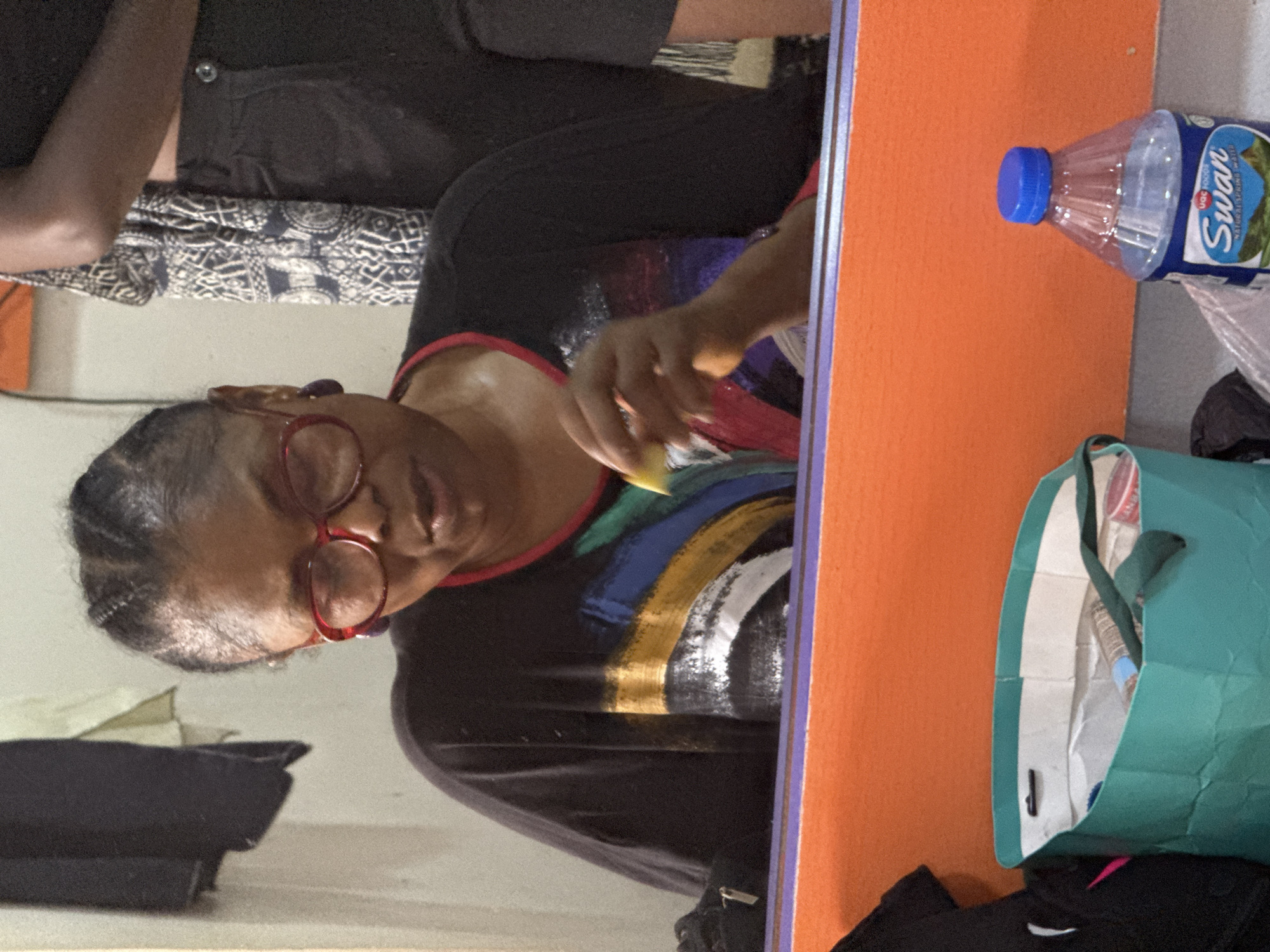 A woman wearing red glasses and a colorful shirt is seated next to an orange surface on which there is a bottle of Swan natural spring water and a green shopping bag. The location appears to be in Abuja, Nigeria, based on the brand of water.