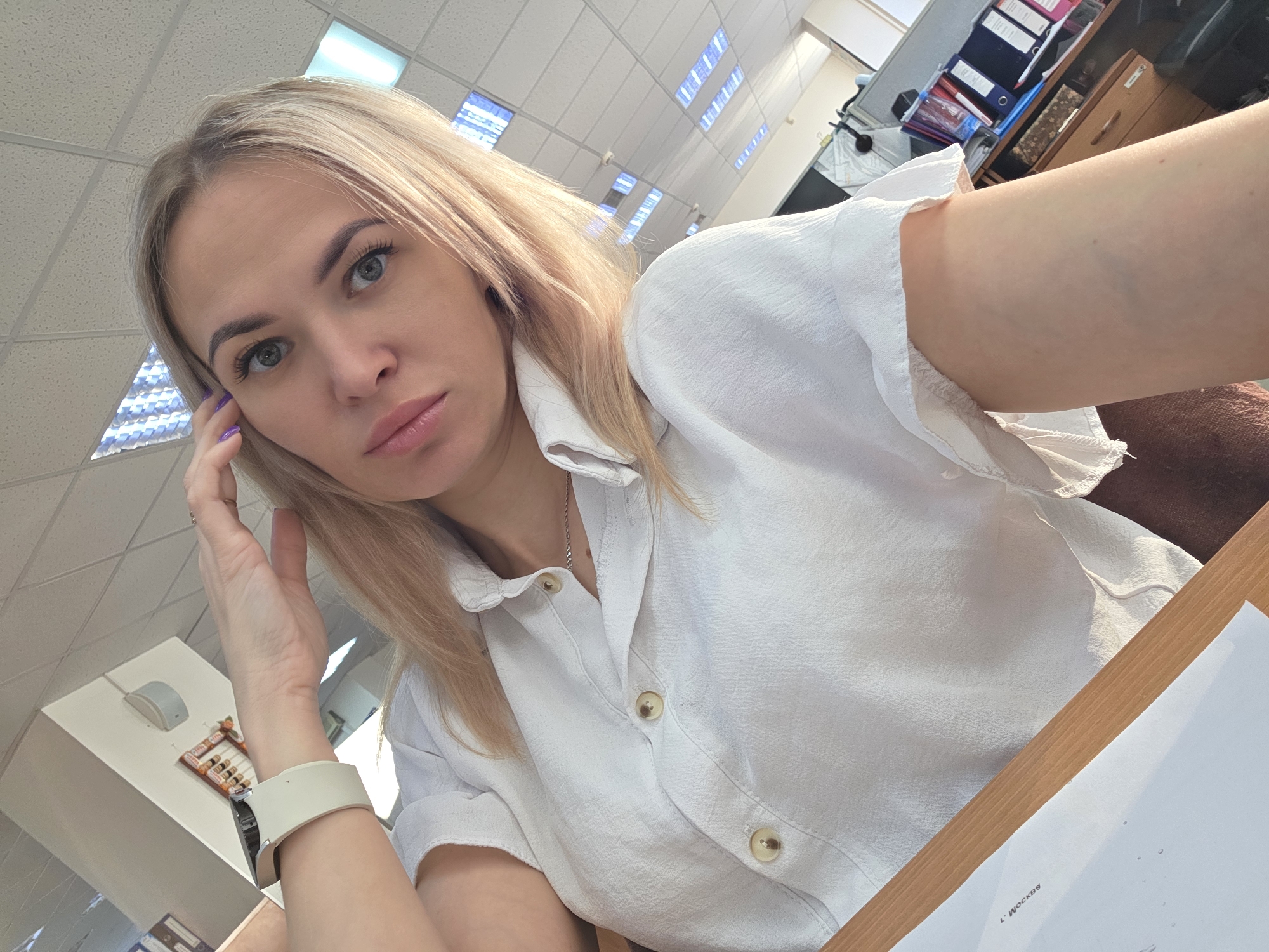 A woman with blonde hair, wearing a white button-down shirt and a white smart watch, is seated at a wooden desk. Her left hand is touching her face near her temple, and she's looking towards the camera. The location appears to be an office environment, evidenced by the fluorescent lighting, files, and office supplies visible in the background. A document is present on the desk in front of her, labeled "L. Moscow" in Russian. The picture was taken in the city of Chaikovsky, Russia.