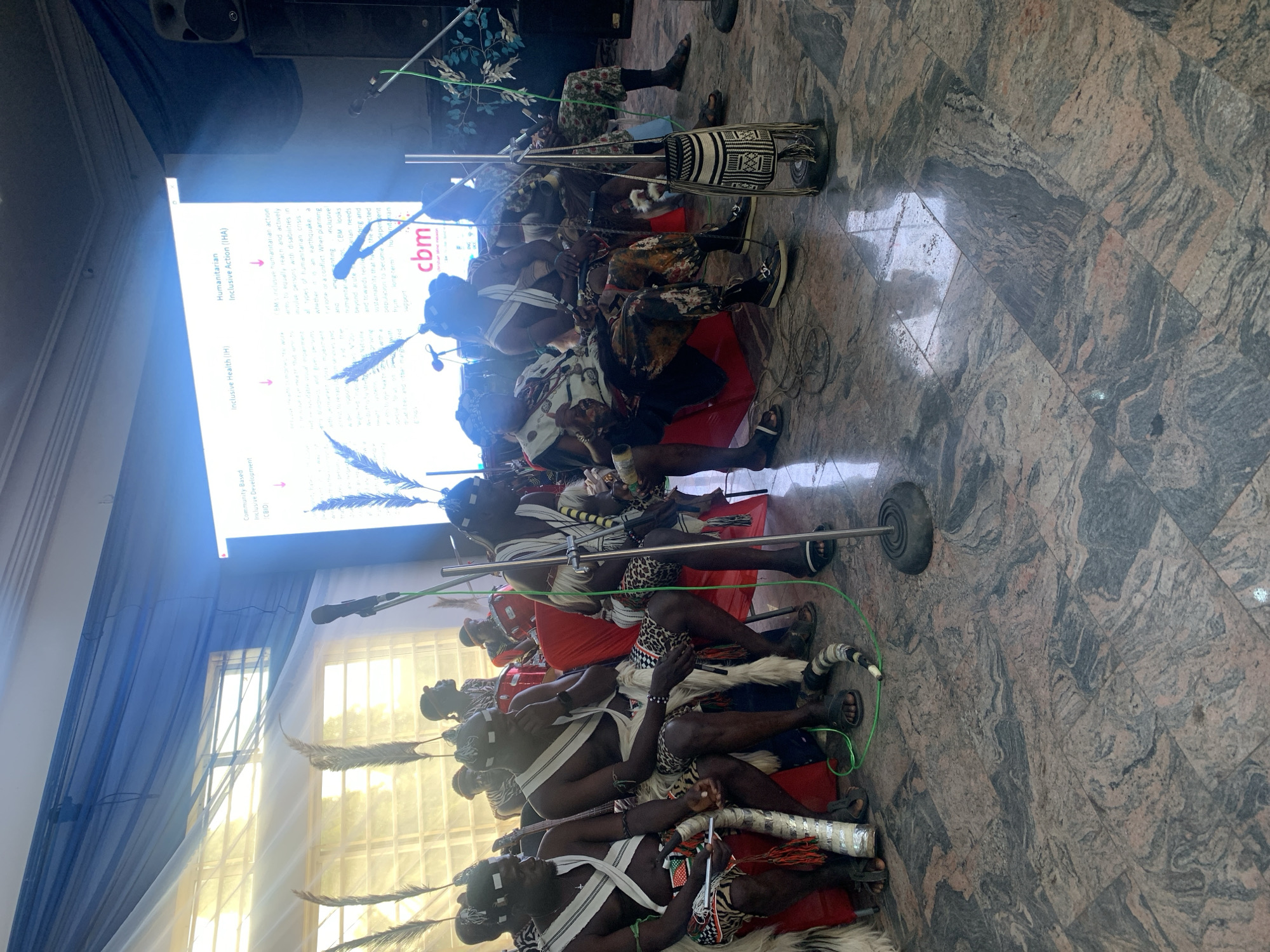 In a large room with a granite tiled floor, a group of men dressed in traditional attire are seated. They wear headbands, white sashes across their chests, and patterned loincloths. Some are adorned with necklaces and wristbands. They are seated on red chairs and are facing a projector screen displaying text and the logo "cbm". The screen is partially obscured by large, feathery decorations. Microphones on stands are positioned in front of some of the men. In the background, other individuals are visible, including one playing a drum. The lighting suggests an indoor event. The scene appears to be taking place in the city of Gura Top, Nigeria.