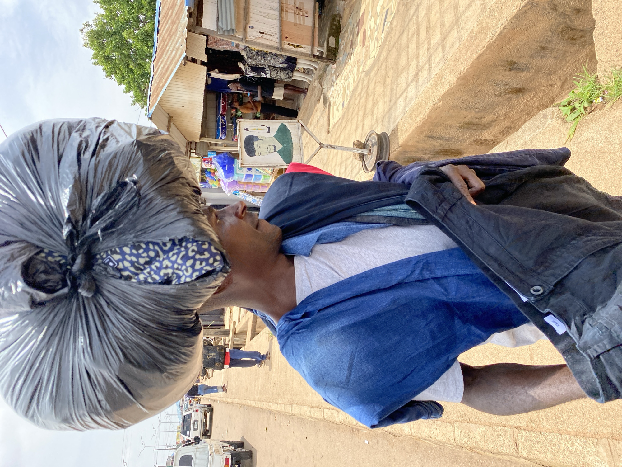 A man in Bukuru, Nigeria, carries a large black plastic bag on his head, balanced by a blue and white patterned fabric. He wears a white t-shirt and a blue cloth draped around his shoulders. In his hands, he holds dark-colored clothing. The man walks along a dirt path next to a street with vehicles, including a white van and a white pickup truck. To his right, a market stall with a corrugated metal roof displays clothing on mannequins and shelves. A sign with a drawing of a man stands near the stall. The environment appears to be an outdoor market on a cloudy day.