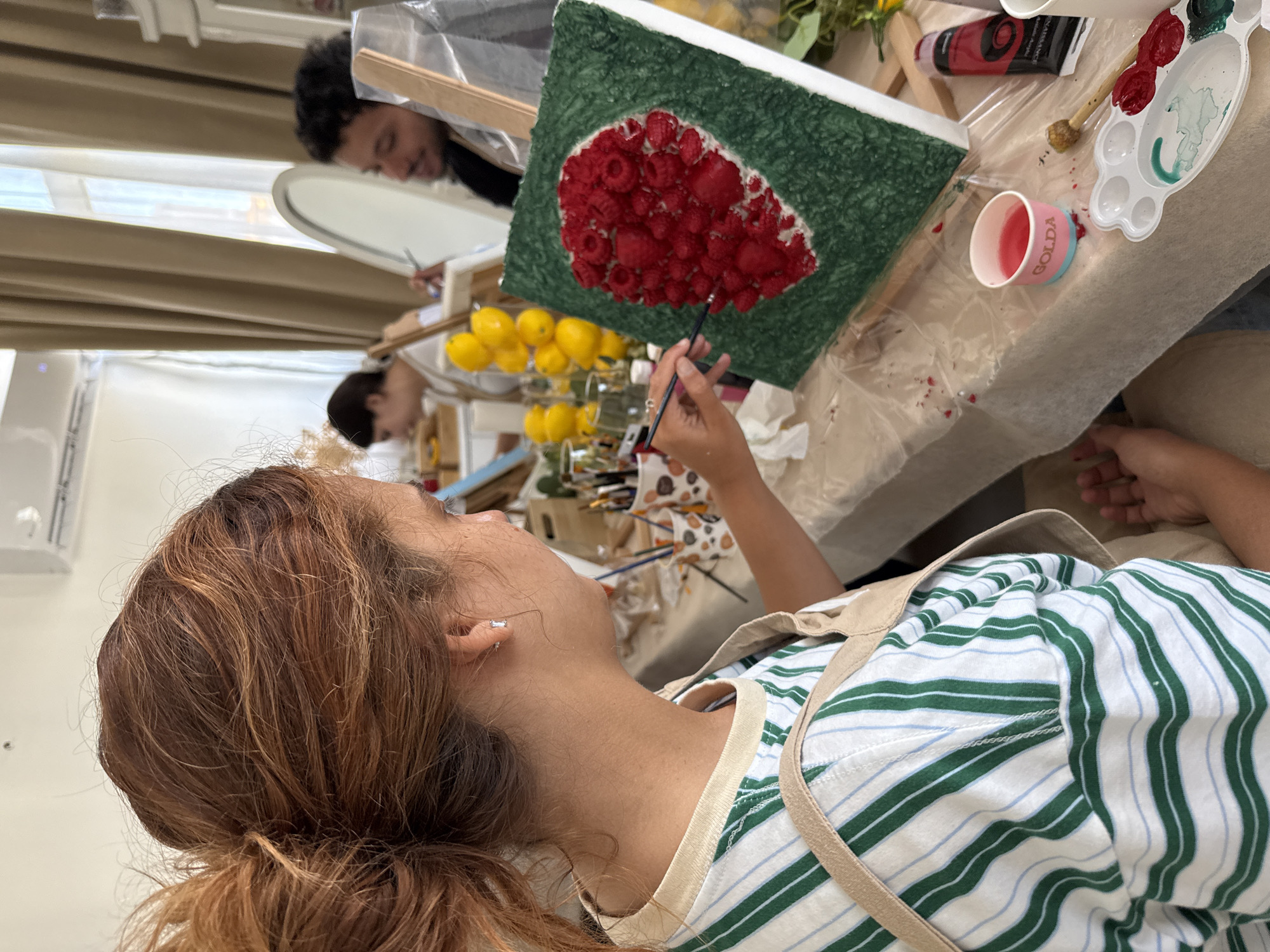 The image depicts an art class, likely a workshop, in what appears to be Tel-Aviv, Israel, based on the name "Golda" visible on a cup, potentially referencing Golda Meir. A woman with auburn hair pulled back into a ponytail, wearing a green and white striped shirt and a beige apron, is painting a textured canvas. The canvas features a green background with a cluster of red, raspberry-like objects in the center. She holds a thin paintbrush in her right hand, applying red paint to the artwork.
On the table, various art supplies are visible, including a paint palette with red and green paint, a tube of red paint, several paintbrushes, and a cup containing a red liquid. A collection of lemons is visible behind the canvas.
In the background, another person is working on a painting, reflected in a mirrored surface. A second individual is partially visible in the background as well, further suggesting an art class setting. The environment is well-lit, indicating it could be a studio or workshop space. The focus is on the woman painting, with the other elements contributing to the setting and context.