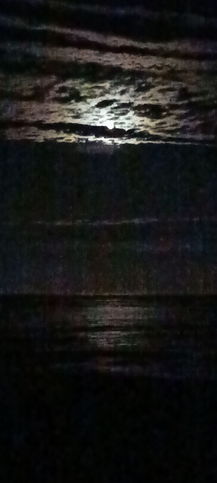 The image is a dimly lit photograph of a body of water under a cloudy night sky with moonlight. The clouds are dark and textured, with patches of bright moonlight filtering through them. The water below is dark and appears calm, with ripples and reflections of the moonlight. The scene is abstract and atmospheric, with a sense of mystery and tranquility. The location is described as "Unknown, Unknown," suggesting a lack of specific geographical information.
