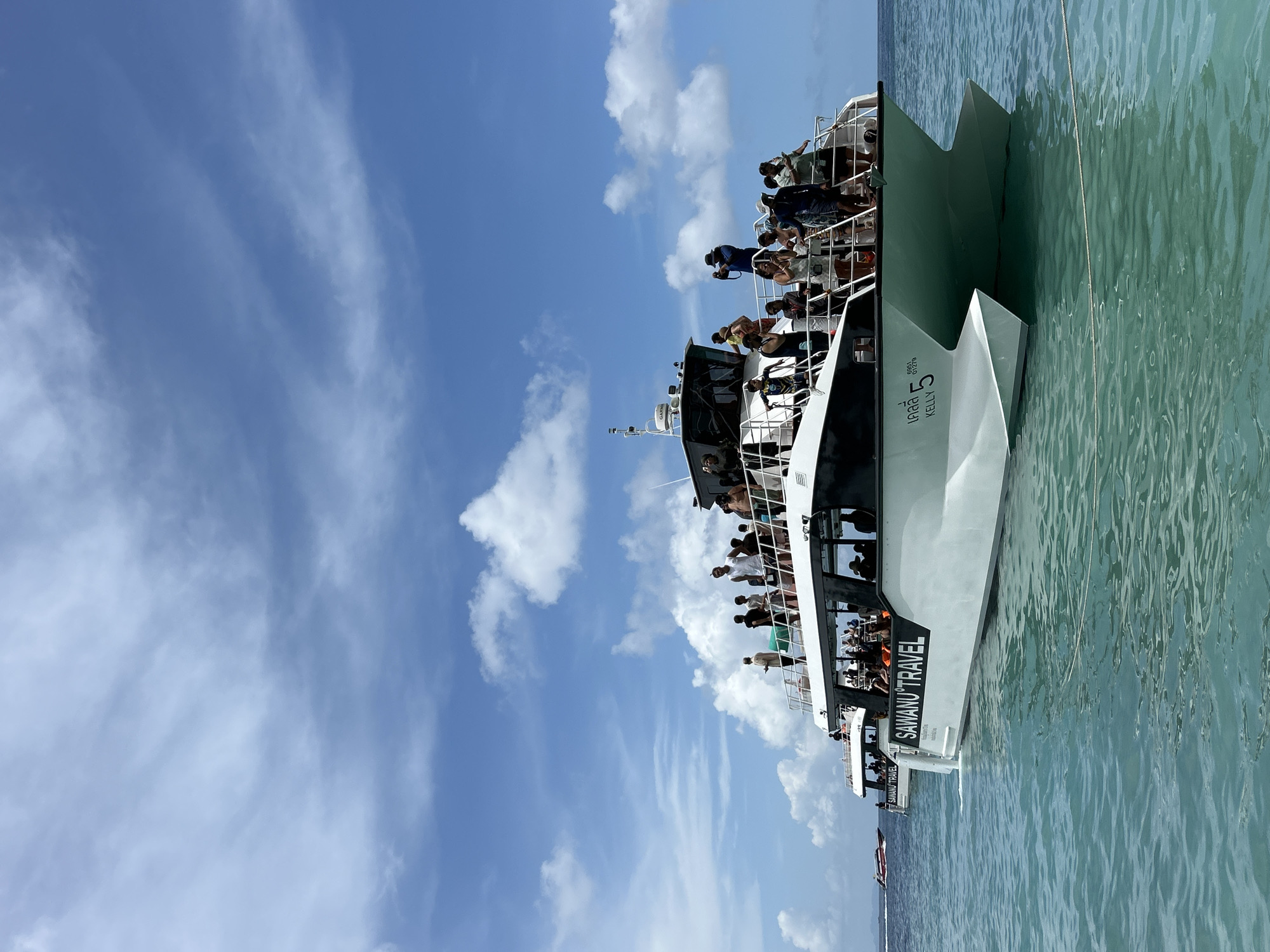 In Ko, Thailand, a white catamaran, "SAWANU TRAVEL KELLY 5," is moored on turquoise waters under a bright blue sky dotted with fluffy white clouds. The boat is filled with passengers standing on its upper deck and peering through the windows of its lower cabin. In the background, another smaller boat speeds across the water, and a distant island is visible on the horizon. The scene captures a typical day for tourists in this tropical paradise, likely enjoying a boat tour or transfer.
