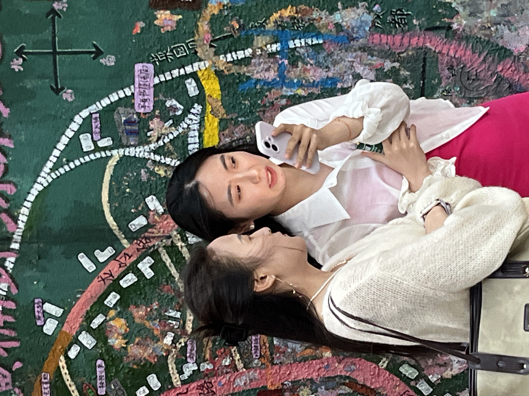 Two women are lying down together on a surface painted with a map of Jiulongpo District in Chongqing, China. One woman is holding a smartphone and appears to be taking a photo or video. The other woman is smiling and looking at her companion. The map includes various features of the district, such as roads, buildings, and green spaces.