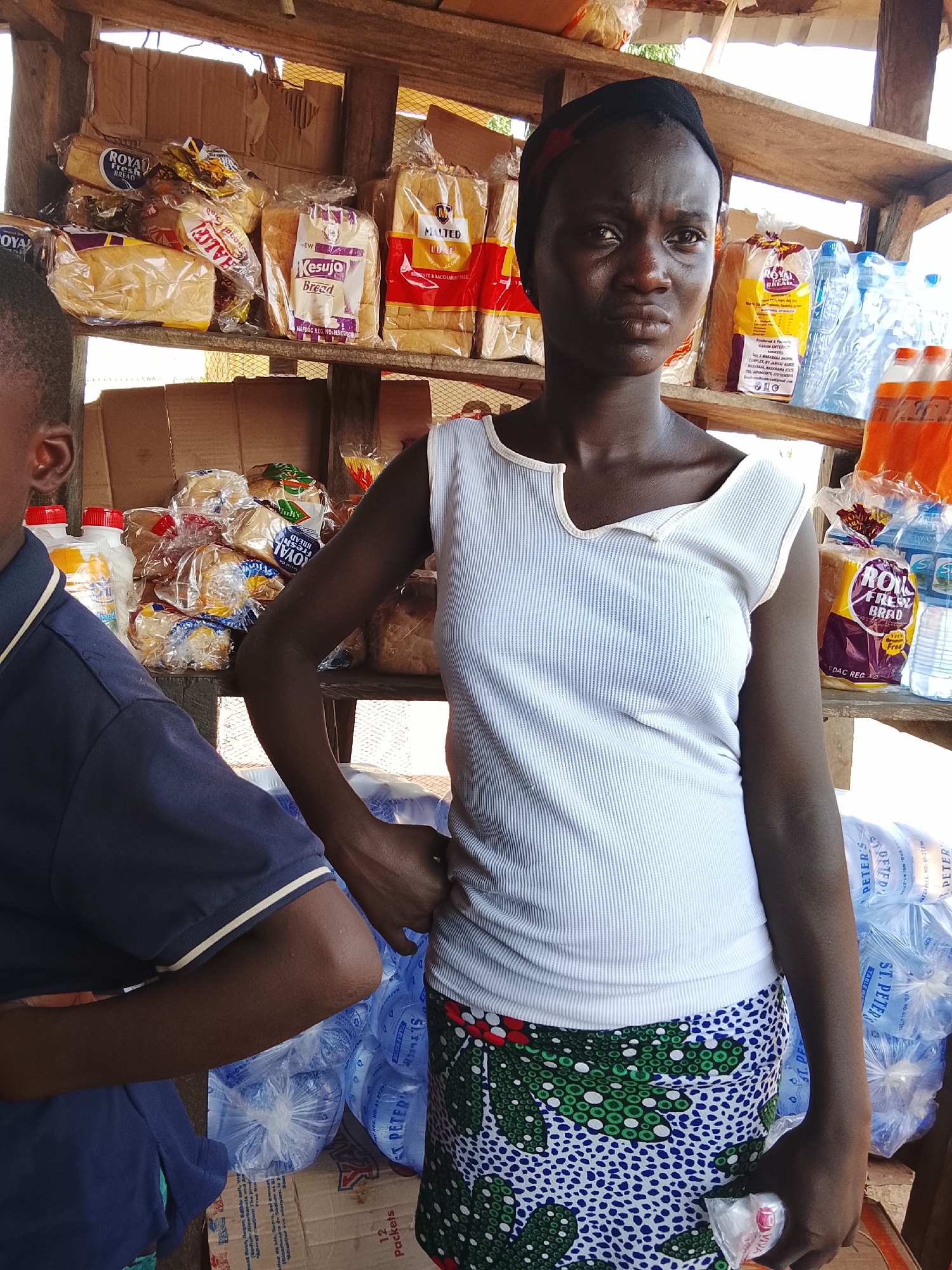 In Akwanga, Nigeria, a woman stands behind a stall filled with food items and bottled water. She is wearing a white ribbed tank top and a patterned skirt. In front of her, partially obscuring her lower body, is another person wearing a navy blue polo shirt. The stall's wooden shelves are stocked with various bread loaves in plastic packaging, including "ROYAL FRESH BREAD" and "MALTED LOAF." Several bottles of water are also visible, as well as bottles of orange-colored beverages. The background suggests an outdoor or semi-outdoor market setting with cardboard boxes and wooden structures.