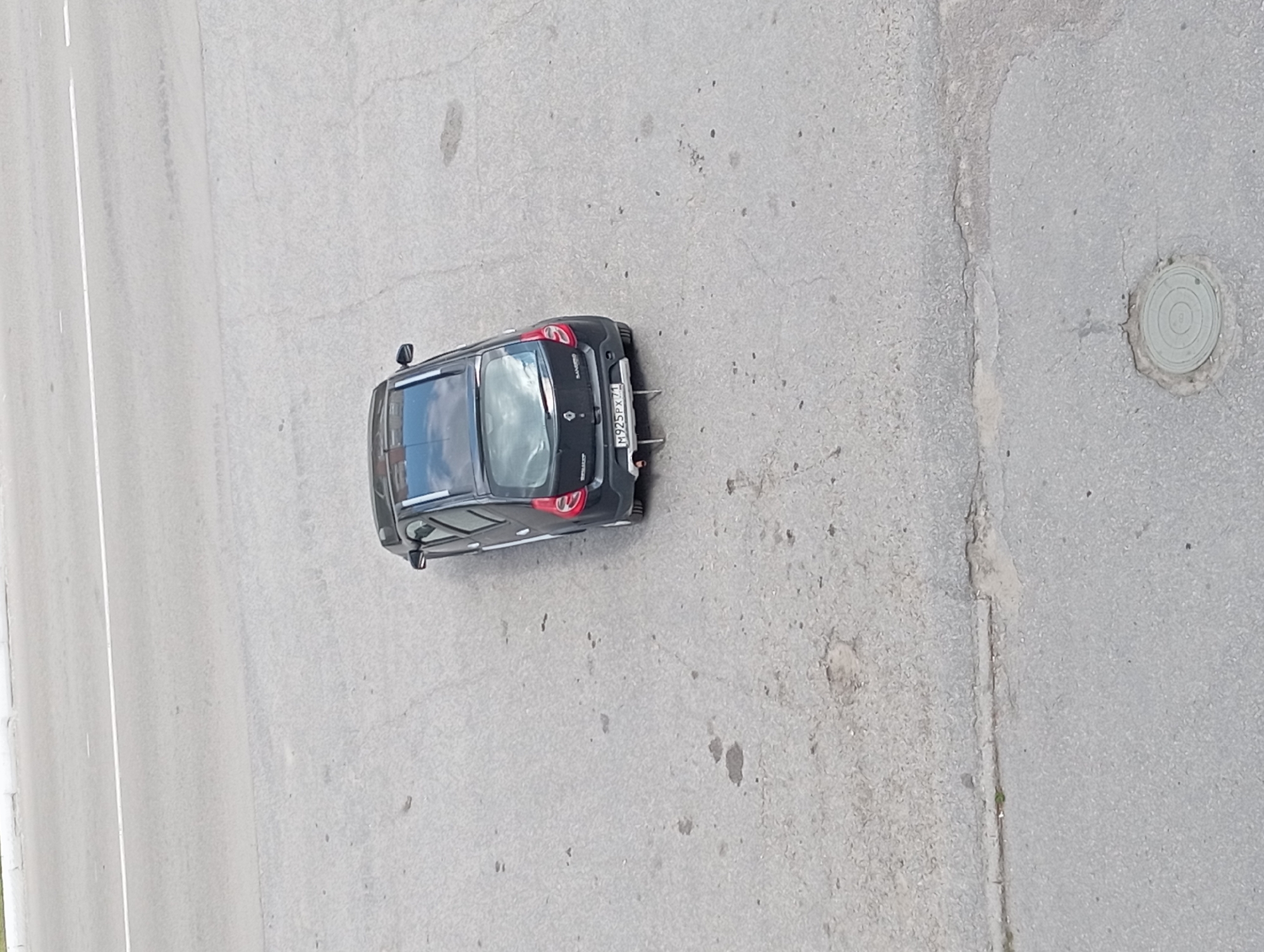 A black Renault Sandero hatchback is parked on a cracked asphalt surface. The car has a license plate that reads "M925PX 71". There is a white line marking a road in the background. The scene appears to be in a city, possibly Suvorov, Russia, given the license plate format. The ground is a dull grey asphalt with dark patches and cracks. A circular manhole cover is visible in the lower right portion of the image. The overall lighting suggests it is daytime.