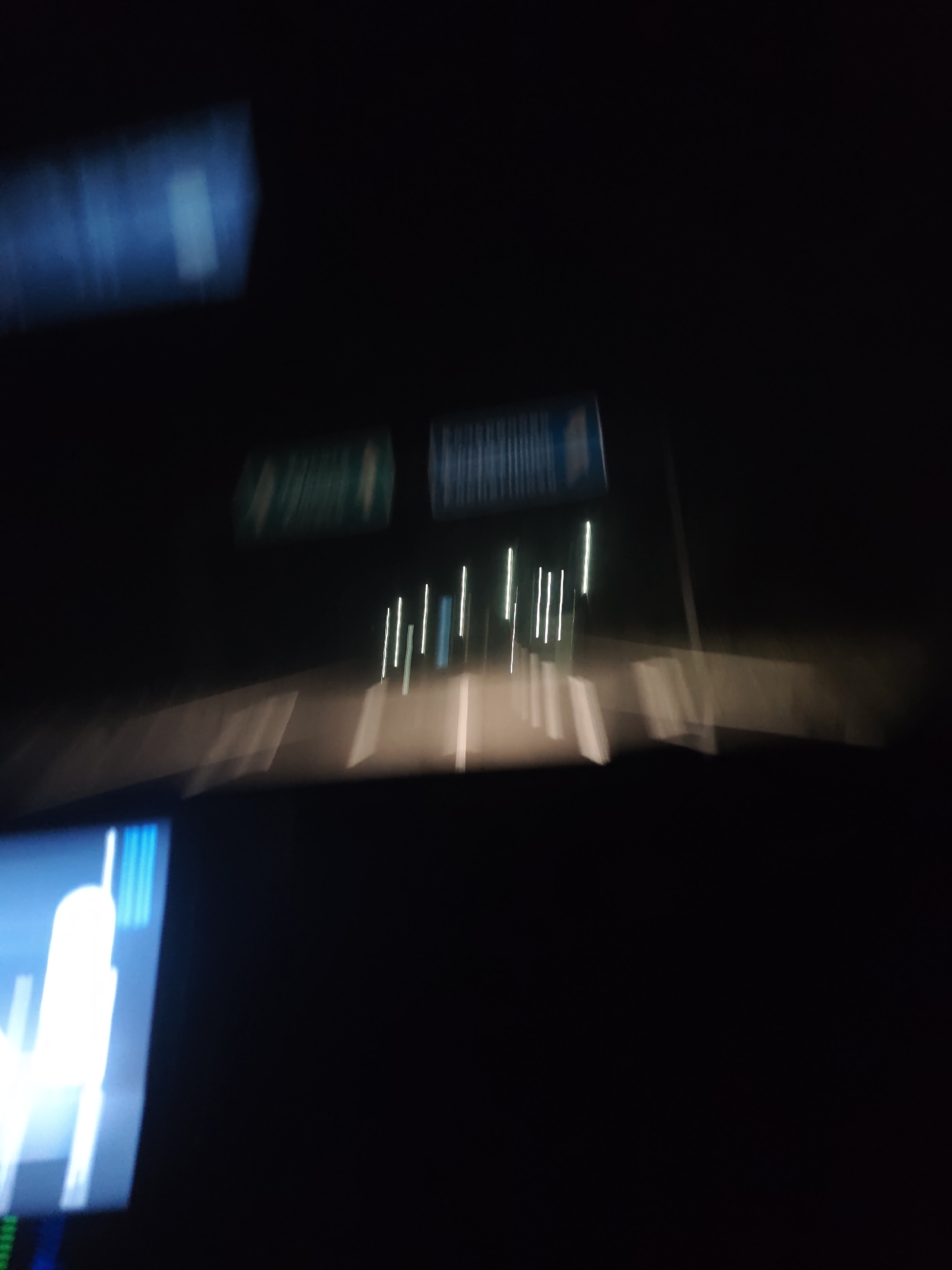 An out-of-focus, nighttime scene taken from inside a moving vehicle. The vehicle is traveling on a highway or well-lit road. Road signs are visible, but illegible due to the blur. There are white lines on the road and streetlights or reflectors on the right side of the road. Inside the vehicle, a dashboard screen is visible on the bottom left of the frame and another blurry object appears at the top left. The image is too blurry to provide further details. There is no evidence that this image is in Barakovo, Bulgaria.