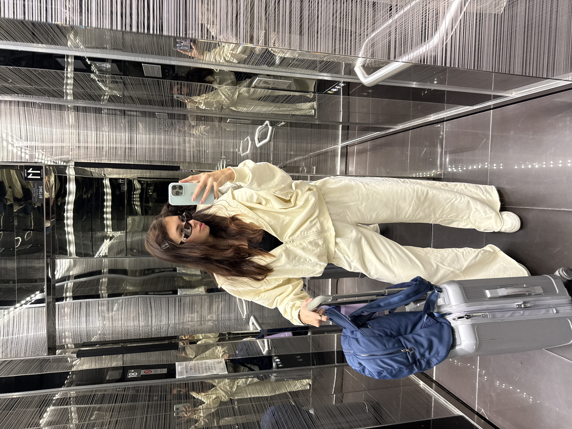 This full-length mirror selfie captures a young woman inside a modern, highly reflective elevator or hallway, likely in Givatayim, Israel. The setting features dark, polished floors and walls made of numerous thin, vertical, metallic or mirrored strips, creating a shimmering, almost abstract effect. Bright artificial lighting illuminates the space.
The primary subject is a woman with long, dark brown hair, wearing dark sunglasses. She is dressed in a casual, cream or off-white colored two-piece tracksuit, consisting of a hooded jacket and matching pants, over a black top. White sneakers are visible on her feet. She is holding a light blue smartphone in her right hand, angled to take the mirror selfie.
Beside her, on the dark floor, rests a silver-colored, hard-shell suitcase with wheels, indicating travel. A dark blue duffel bag or backpack is placed on top of the suitcase. A white, minimalist handrail is visible on the right side of the mirrored wall.
The activity taking place is the woman posing for and taking a mirror selfie. Her expression, partially obscured by sunglasses, appears neutral and posed.
Notable details include reflections of the woman and the interior stretching into the distance. Visible text on signs, albeit mirrored and partially obscured, includes Hebrew characters, with "18" and "ו/או" (and/or) discernable, along with a symbol resembling a prohibition sign. This visible Hebrew text reinforces the provided location context of Givatayim, Israel. The overall impression is one of contemporary design and travel.