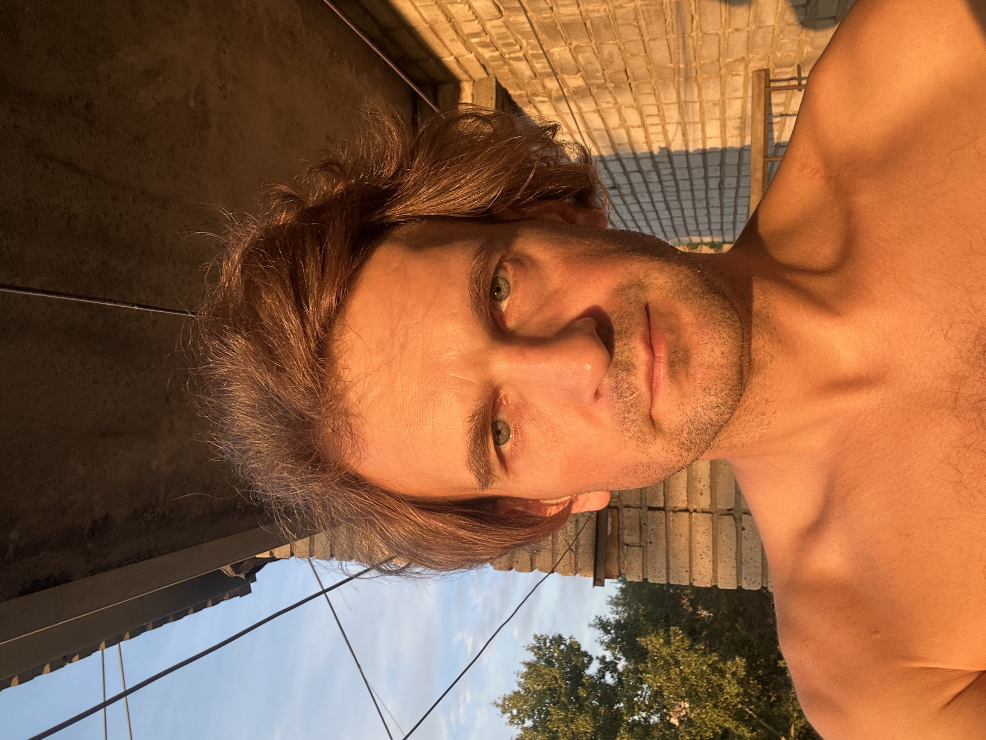 A shirtless man with tousled brown hair and green eyes is photographed from the chest up, outdoors in what appears to be a city environment. The man's face is illuminated by the golden light of what seems to be a sunset. He has a neutral expression and stubble on his chin and upper lip. Behind him, a brick wall is visible, bathed in the same warm light, with a shadow cast upon it. Above the wall, a balcony overhang with wires running across it can be seen. To the left of the man, a green tree and a clear blue sky with scattered clouds are visible. The presence of the brick wall, balcony, and tree suggests an urban setting, possibly residential. Without further information, pinpointing the exact location within Biysk, Russia, is not possible from the visual data alone.