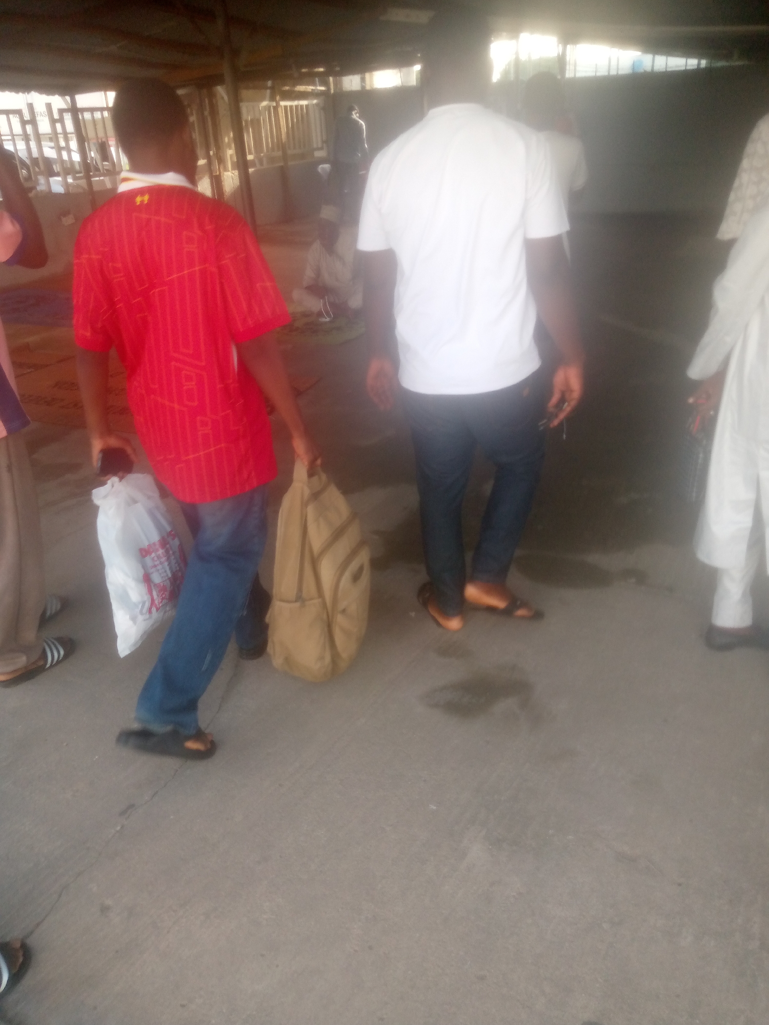 The image depicts a group of people in what appears to be an outdoor market or gathering space in Jalingo, Nigeria. The scene is illuminated by natural light, suggesting it is daytime, possibly late morning or afternoon. The ground is a concrete surface, and there are structures with corrugated metal roofing visible in the background, hinting at an open-air market or a shaded gathering area.
The primary subjects are several individuals, seen from the waist down and from behind. In the foreground, a person wearing a red t-shirt with a yellow insignia and blue jeans is holding a white plastic bag in their left hand and a tan backpack in their right. To their right, another person is walking away, wearing a white t-shirt and dark trousers, also holding keys in their right hand. Further to the right, a third person is visible, wearing white traditional attire. In the mid-ground, other figures are present, some seated.
The activity appears to be people moving through or congregating in the area, possibly after a religious service or during a market day. The atmosphere seems casual. No explicit text is clearly legible in the image, though the red t-shirt has a pattern that might include some text or logos, but it is not discernible. The overall impression is of a common day in a Nigerian community.