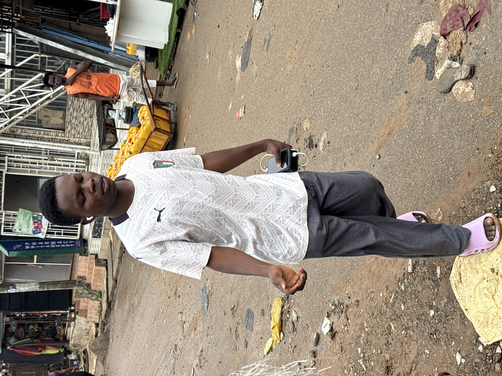 The image depicts a street scene in Abuja, Nigeria, with a young Black male walking towards the viewer. He is wearing a white t-shirt with a geometric pattern and an Italian flag emblem, dark gray sweatpants, and light pink slides. He has white earbuds in his ears and is holding a smartphone and a portable charger in his right hand. The ground is a wet, muddy dirt road with scattered debris, including plastic bags and what appears to be a discarded shoe. In the background, there are buildings with shops, including a pharmacy with signage. Another young Black male in an orange tank top and white shorts is visible in the middle ground, standing near a cart loaded with yellow jerrycans. The overall impression is of a busy, somewhat cluttered urban environment.