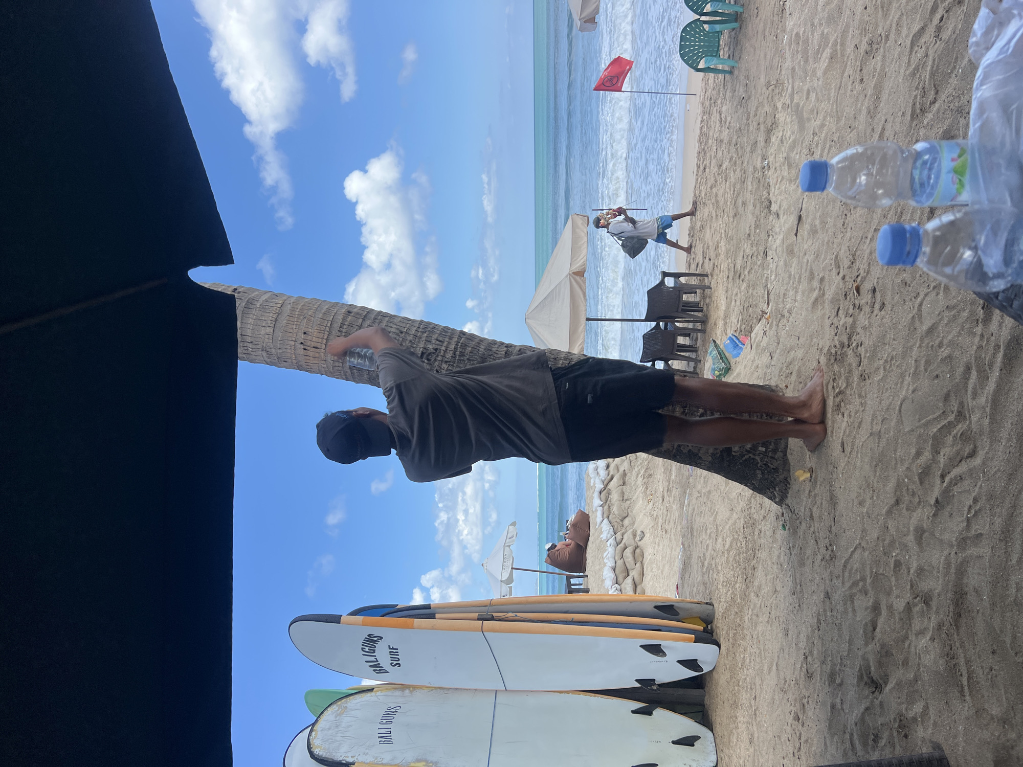The image depicts a beach scene in Kuta, Indonesia, with a man standing next to a palm tree, holding a bottle of water and appearing to be affixing it to the tree. In the foreground, several surfboards are propped up, with "BALIGUNS SURF" visible on two of them. The background shows the ocean with gentle waves, a person relaxing on a beanbag chair under a beach umbrella, and another person walking along the shore carrying a bag and what appears to be a souvenir. The sand is the primary ground surface, with some scattered plastic bags and bottles. A red flag is planted in the sand near the water. The sky is blue with scattered white clouds.