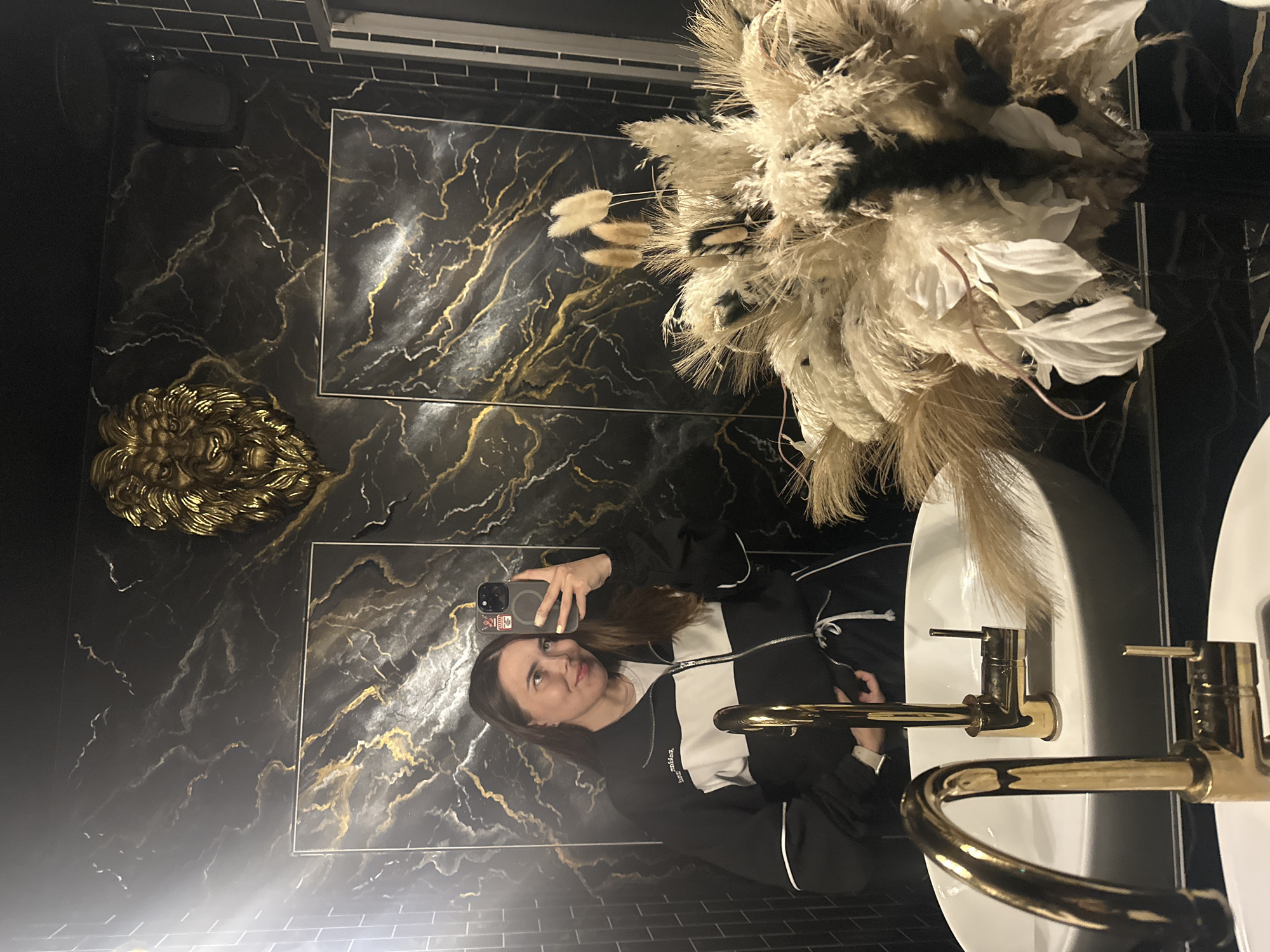 The image depicts a woman taking a selfie in a bathroom, likely located in Perm, Russia. The bathroom features black marble walls with gold veins, a gold lion head sculpture mounted on the wall, and black subway tiles. In the foreground, two gold faucets are visible over a white sink. To the right, a large bouquet of dried pampas grass and other decorative elements sits in a vase. The woman is wearing a black and white tracksuit and is holding a smartphone to take the photo. Her reflection is visible in the mirror, showing her upper body and face.