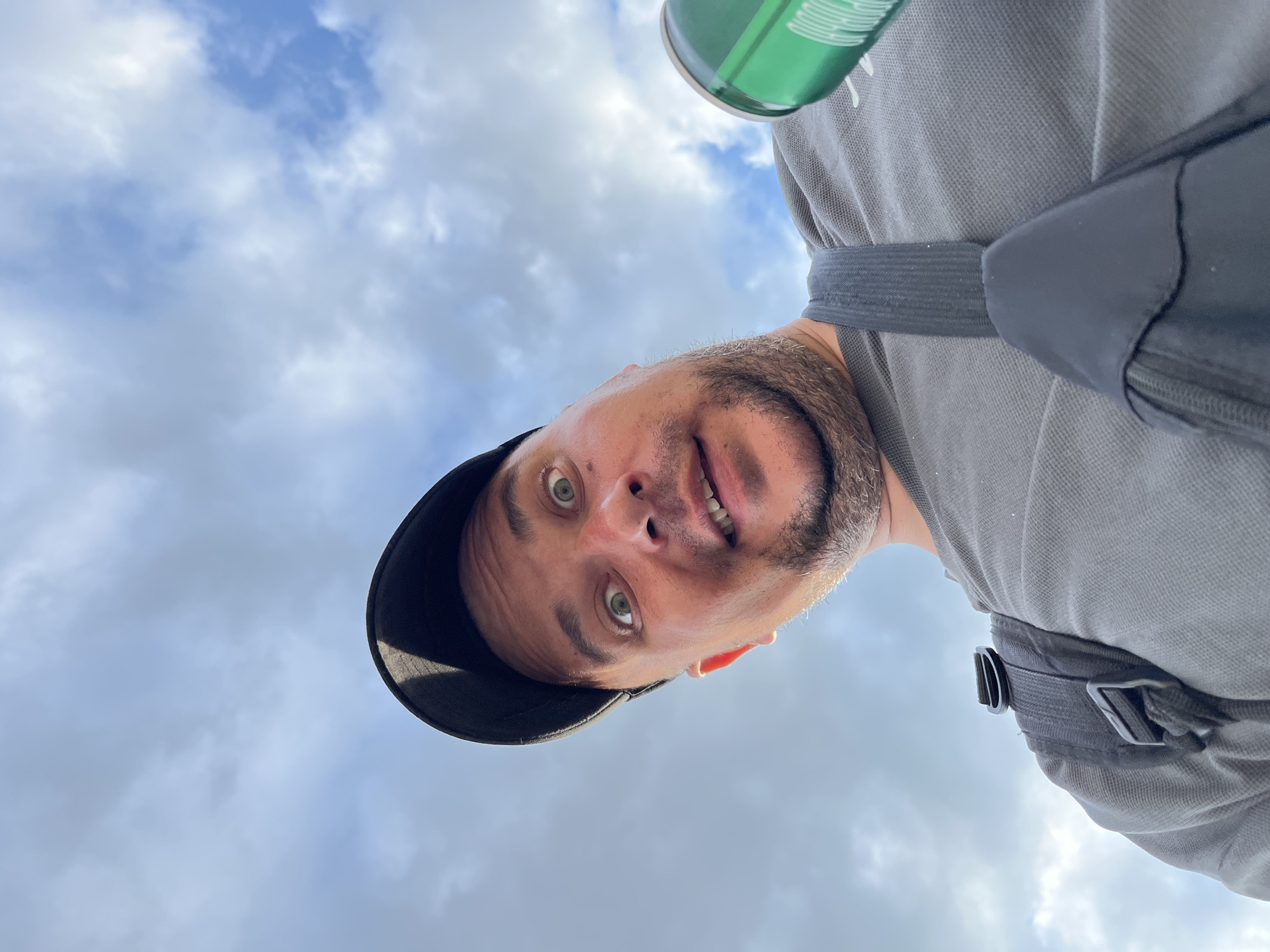 A white male, appearing to be in his late 30s to 50s, is captured in a low-angle, close-up view from the chest up. He has a short beard or stubble and light-colored, wide-open eyes, with a slightly open mouth, conveying a surprised or engaged expression. He wears a black baseball cap, a grey short-sleeved polo shirt, and a black sling bag or small backpack with a visible strap across his chest and shoulder. In his right hand, partially visible on the right side of the frame, he holds a green metallic can. The background consists entirely of an overcast sky with scattered clouds. The image does not contain any specific landmarks or features that would allow for the identification of a precise location within Minsk, Belarus.