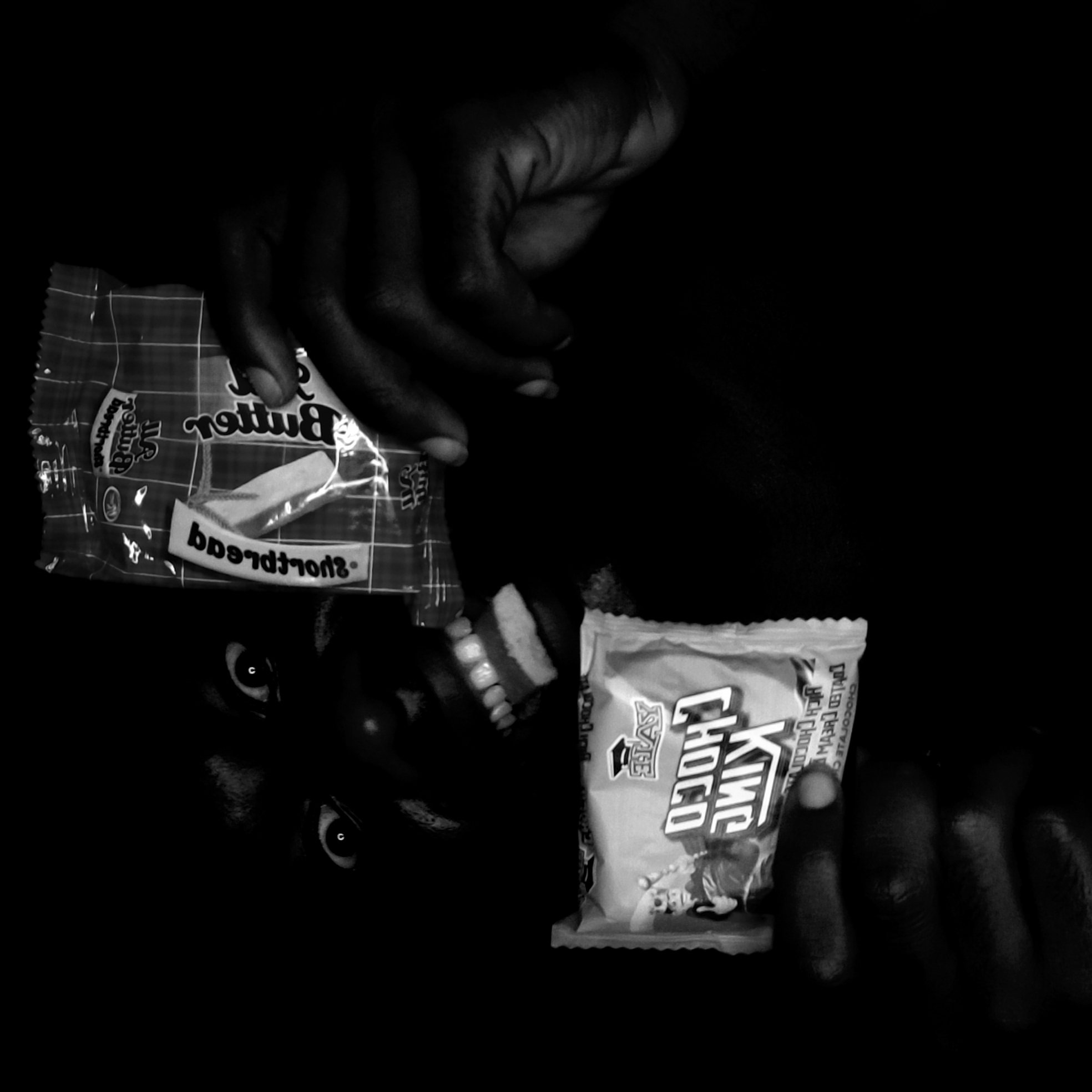 The image is a black and white photograph of a person with dark skin holding two packages of candy near their face. One package is labeled "Butter Sports" and the other "King Choco." The person is looking upward toward the "Butter Sports" candy. The background is dark and obscures the environment. The location of Narayi, Nigeria cannot be verified from the image.