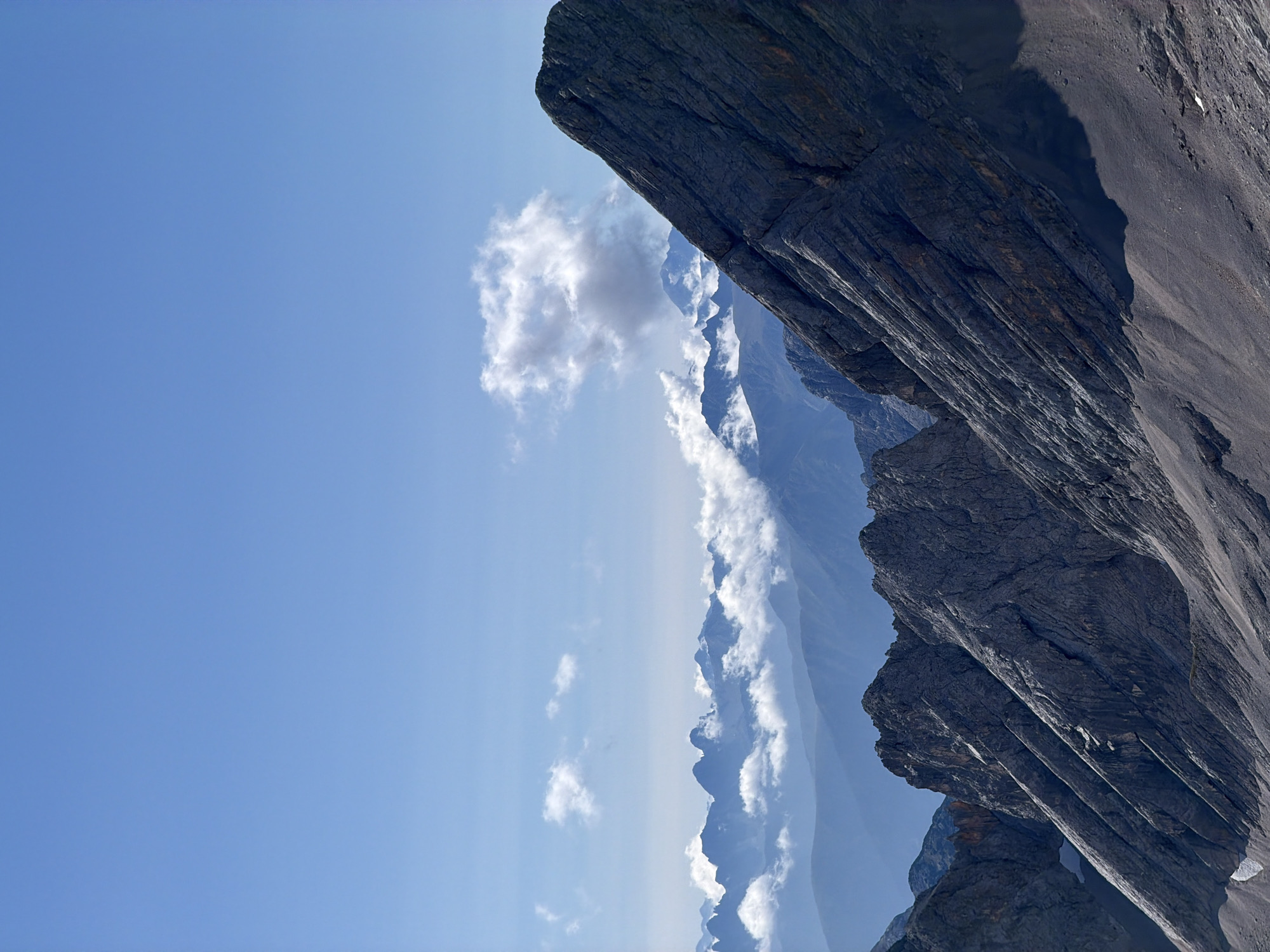 The image displays a dramatic mountain landscape under a clear blue sky. In the foreground, a steep, rocky slope dominates the right side of the frame, characterized by dark, textured rock faces with visible striations. To the left, jagged mountain peaks emerge, some partially obscured by wisps of white clouds. Further in the distance, layers of mountain ranges recede into a hazy blue, creating a sense of depth and vastness. A few scattered clouds drift in the upper portion of the sky, with one larger, more defined cloud formation to the right. The scene captures the rugged beauty of a high-altitude environment. This is the Zugspitze, Germany's highest peak, located near Garmisch-Partenkirchen.