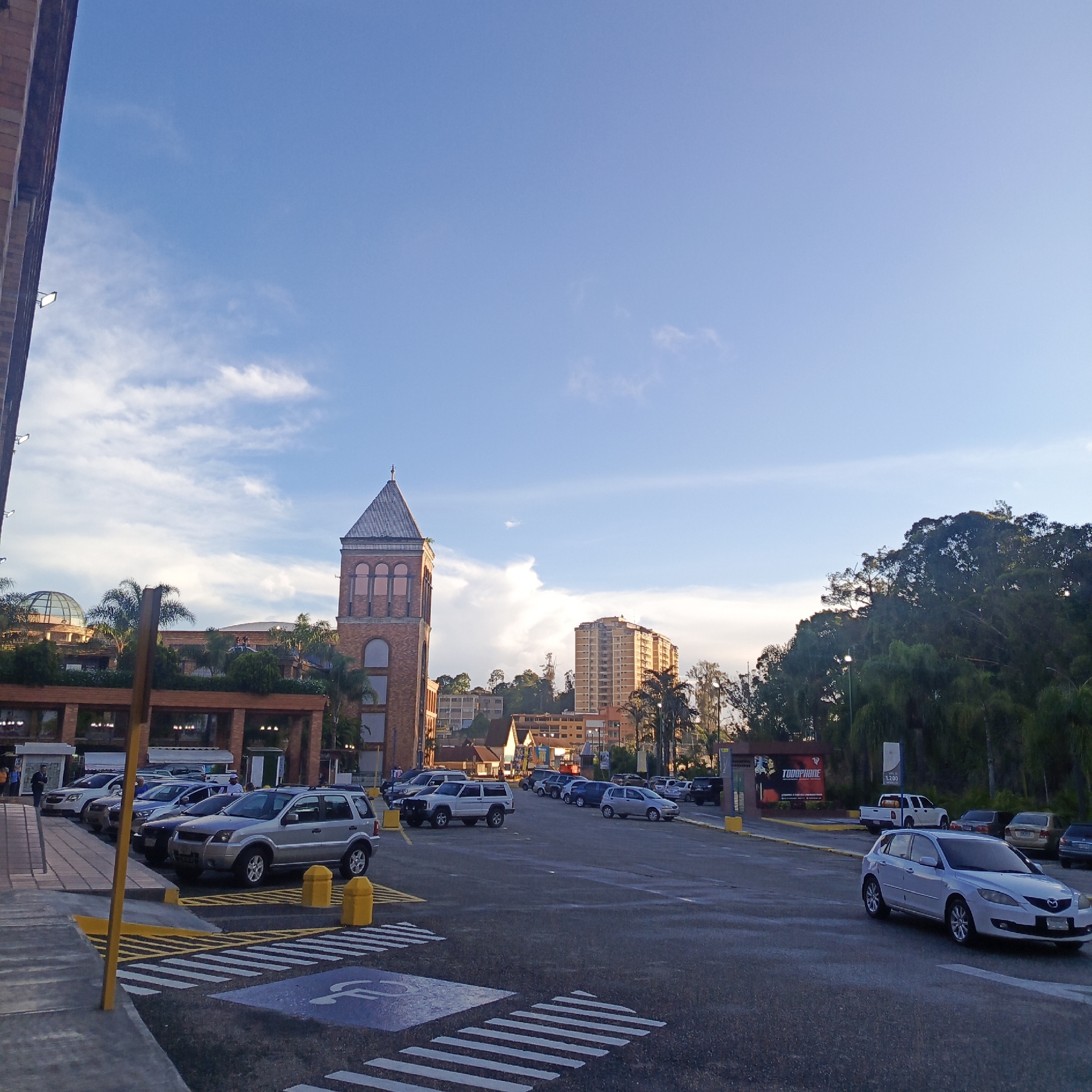 The image depicts a parking lot outside a commercial building in Carrizal, Venezuela. The architecture of the building features a prominent brick clock tower and a domed structure. The parking lot is occupied by several vehicles, including SUVs, sedans, and a white pickup truck. Pedestrian crosswalks and accessibility markings are visible on the asphalt. The sky is blue with scattered clouds, suggesting it is daytime. Trees and other buildings are visible in the background, indicating an urban or suburban environment. The overall scene is a typical view of a commercial area with adequate parking facilities.