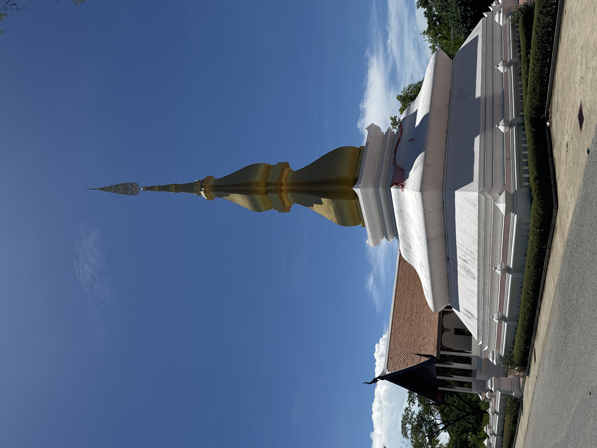 The image depicts a pagoda at Wat Asokaram in Phraek Sa Subdistrict, Thailand. The pagoda features a golden spire, a white base, and a traditional Thai architectural style. The sky is blue with white clouds, and the ground is paved with greenery. The image is rotated.