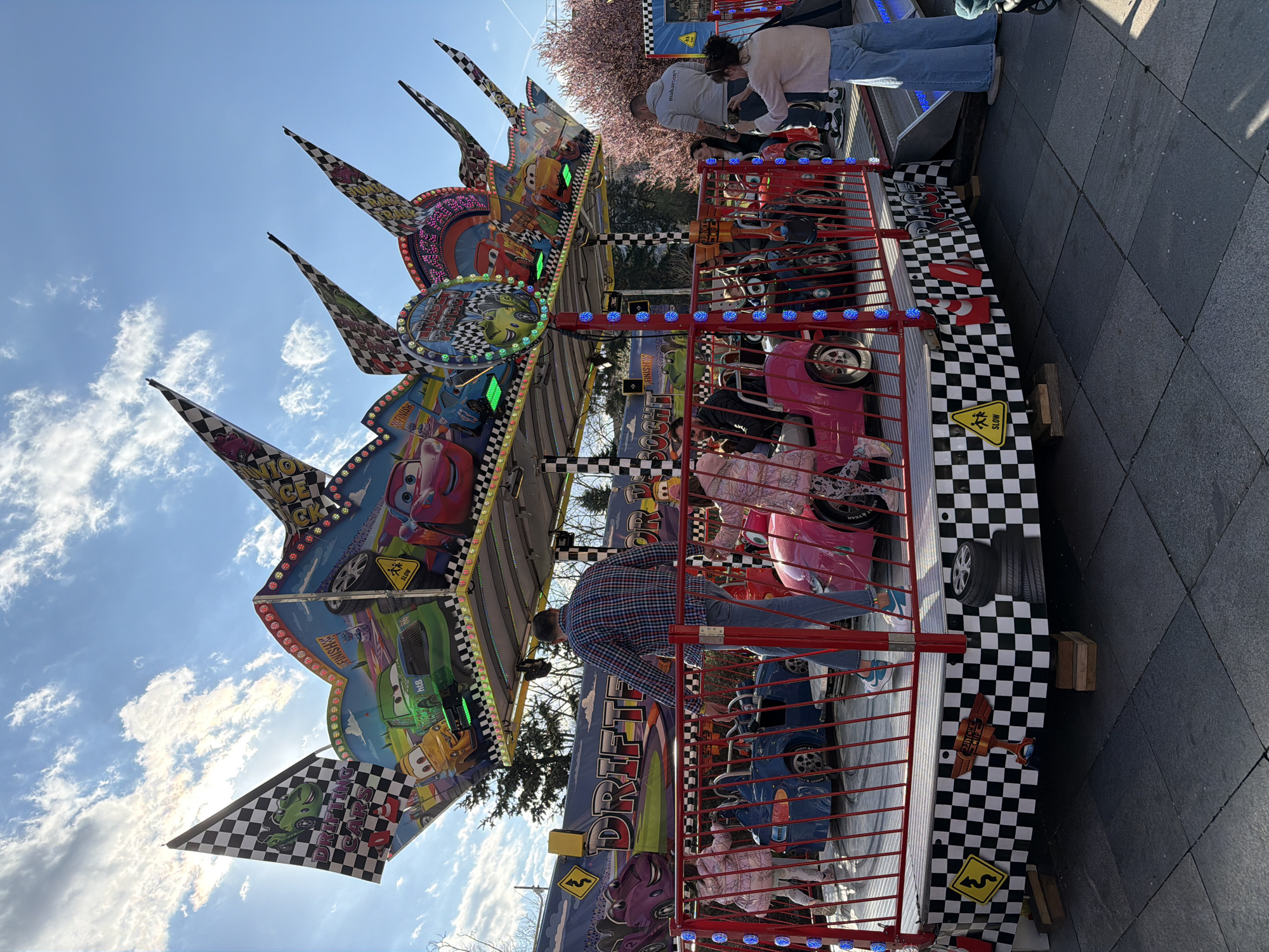 The image shows a children's ride themed around racing cars at an amusement park or fair. The ride is decorated with imagery of cars and checkered flag patterns. People are either riding or waiting near the ride. The ground is tiled. The sky is blue with scattered clouds. Trees are visible in the background. The image was taken in Sofia, Bulgaria.