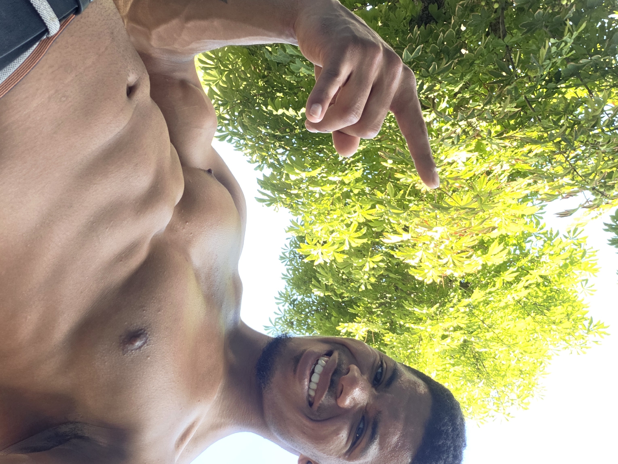 A low-angle, outdoor, close-up selfie taken from below shows a shirtless man with dark skin and short dark hair. He is smiling broadly and looking directly at the camera, with his head tilted. His left arm is bent, and his index finger is extended upward, as if pointing. A portion of his muscular torso is visible, including his chest and abdomen. In the background, a large, vibrant green tree with bright green leaves is prominently featured, bathed in sunlight. The sky is bright and white, suggesting a clear day. The image was taken at the Sea Garden in Varna, Bulgaria.