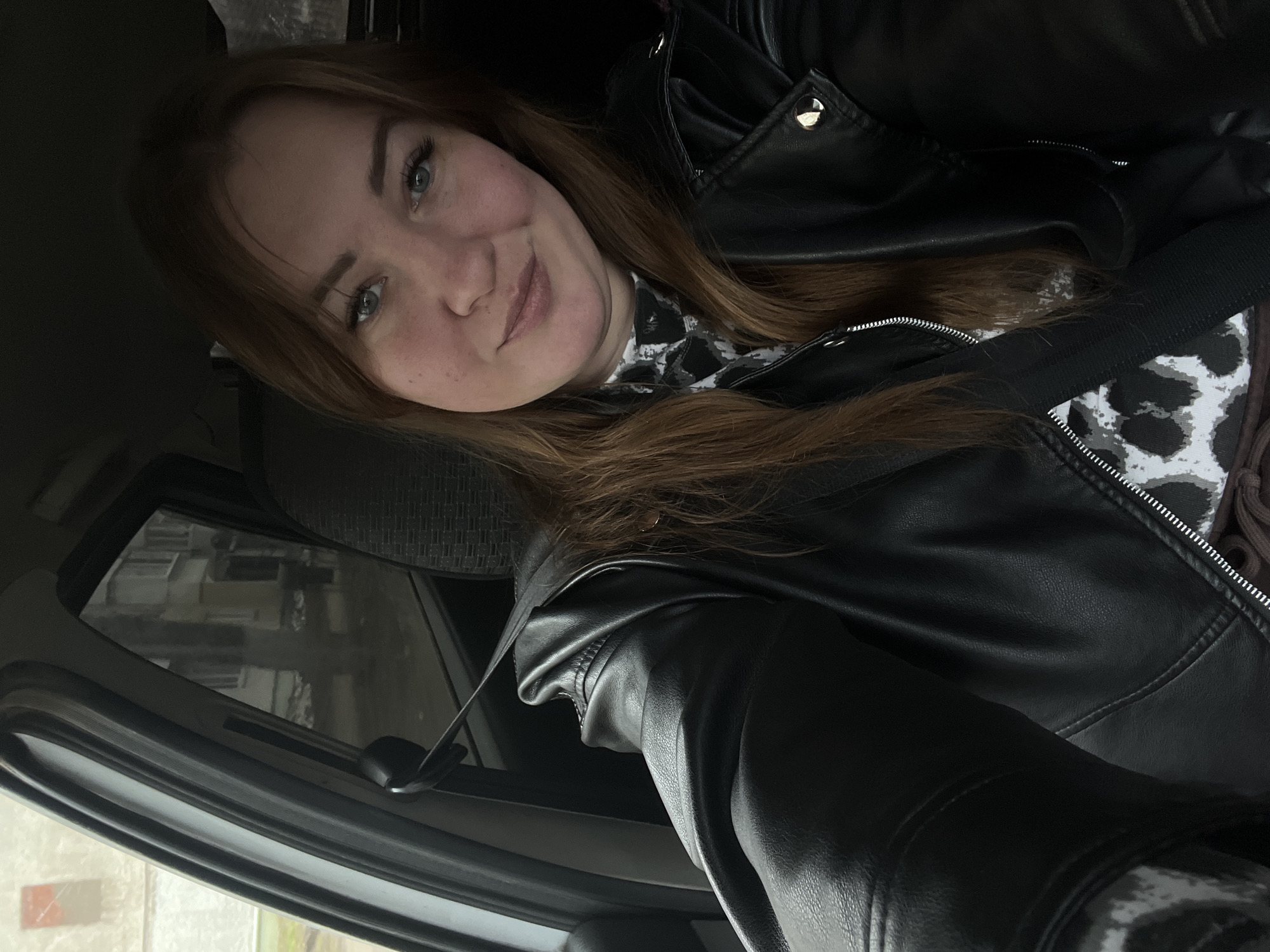 A woman with light skin, reddish-brown hair, and light-colored eyes is sitting in a car, wearing a black leather jacket over a white and black printed top. A black seatbelt crosses her chest. The interior of the car is visible around her. Through the car window, buildings are visible outside, reflecting the city environment. The location is Chaikovsky, Russia.