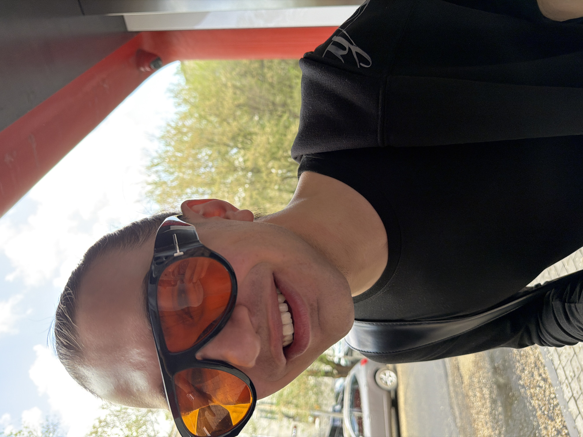 A close-up, slightly upward-angled portrait shows a light-skinned man with short hair and sunglasses standing outdoors. The lenses of the sunglasses have a deep orange hue and reflect the surrounding environment. He is smiling, revealing his teeth. The man is wearing a black long-sleeved shirt with a logo embroidered on the shoulder. A black leather bag strap is visible across his chest. Behind him are trees with green foliage and a blue sky with scattered clouds. A red metallic structure is visible to the left of the man's head. A car parked on the street and a stone-paved walkway is visible on the right side. While the image does not definitively confirm that it was taken in Yerevan, Armenia, it suggests an urban environment with typical city features.