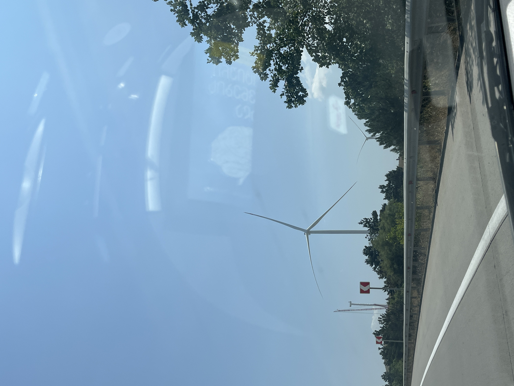 The image shows a bright, clear sky with some reflections on a surface, possibly a windshield. In the background, there are several wind turbines, one of which is prominently displayed with its blades spread. A construction crane is visible to the left of the wind turbine. A highway guardrail runs horizontally across the middle ground, with road signs indicating a curve to the left. Trees and vegetation line the side of the highway. The scene appears to be a rural or semi-rural area in Georgia. The specific location of Ruisi, Georgia, is not directly evident from the visual information in the image itself.
