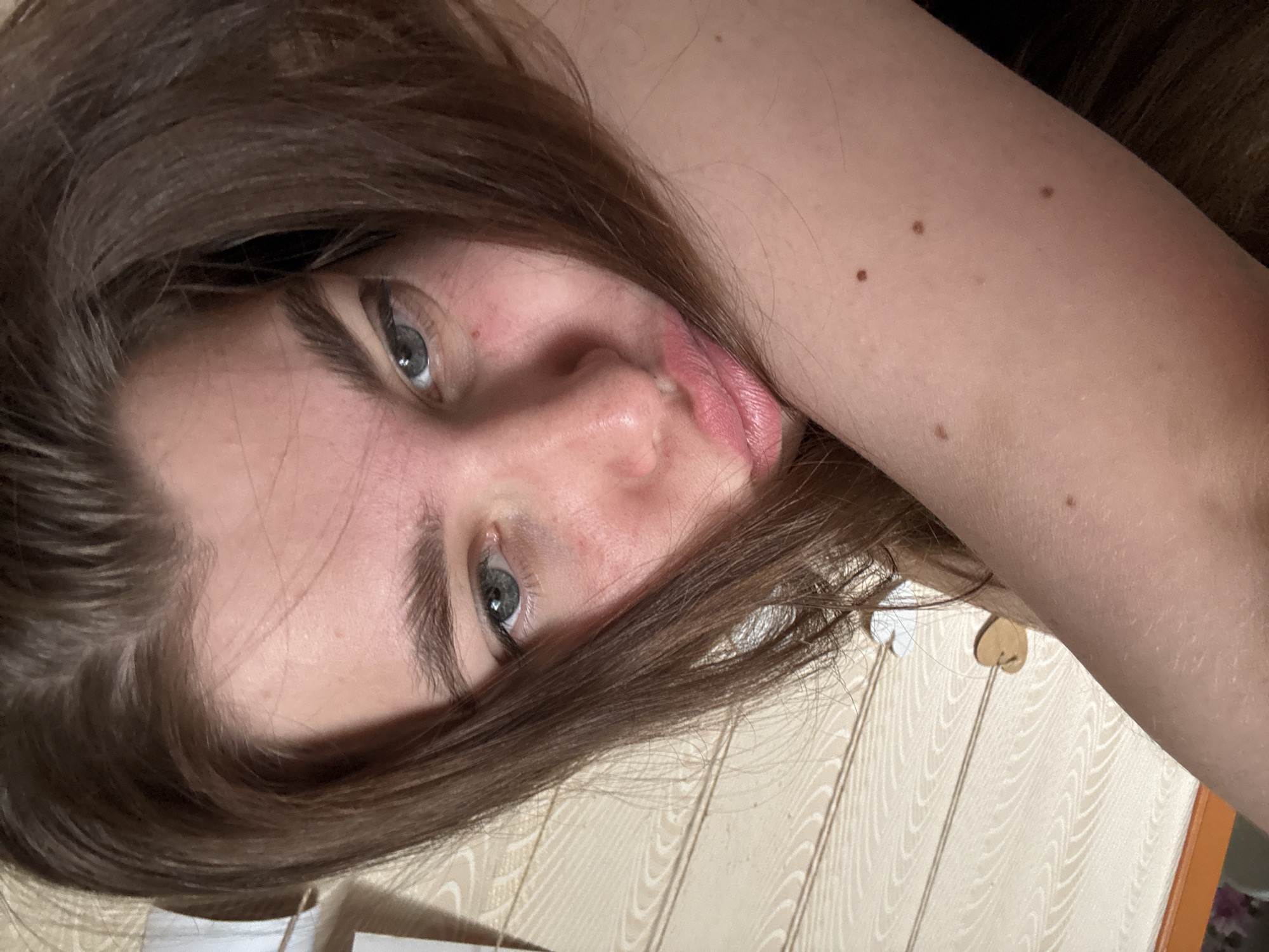 A selfie shot of a fair-skinned woman with brown hair and grey eyes. She is positioned at an angle, with her head tilted to the right. Her lips are slightly puckered. Her right arm is visible, showing some moles on the skin. The background appears to be a light-colored wall with a patterned design, possibly wood paneling. A small wooden heart decoration is visible. There is no evidence to suggest this photo was taken in Мещёры, Russia.