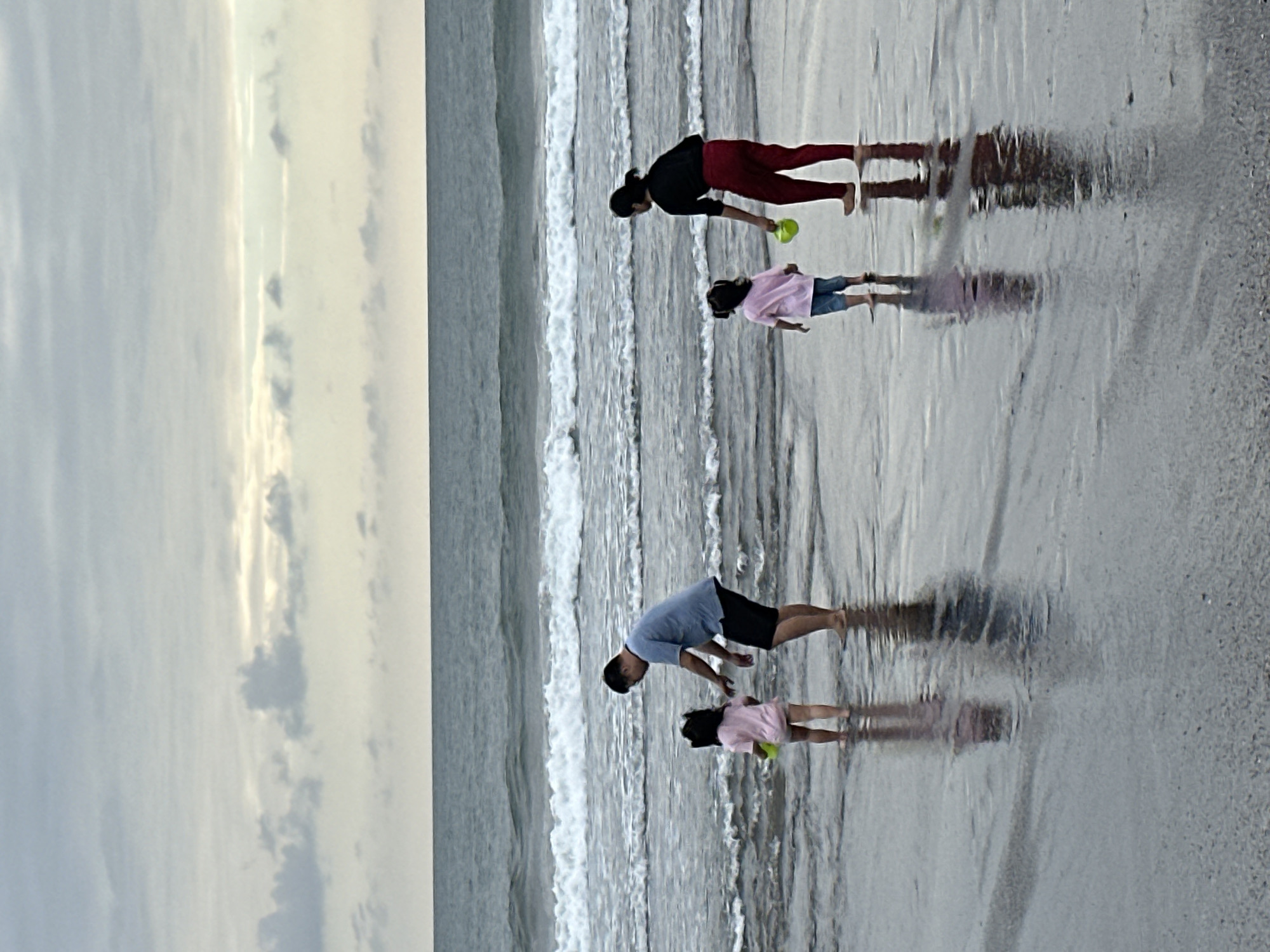 In North Kuta, Indonesia, a family enjoys a day at the beach. The scene captures the vast ocean under a cloudy sky, with gentle waves lapping at the shore. A father, dressed in a blue t-shirt and black shorts, stands in the shallow water with his young daughter, who wears a pink shirt and shorts. He appears to be interacting with her as she holds a yellow toy. Nearby, a woman in a black top and red pants stands with another young girl, also in a pink shirt and jeans, holding a green toy. The wet sand reflects the figures and the sky, creating a serene and picturesque setting.
