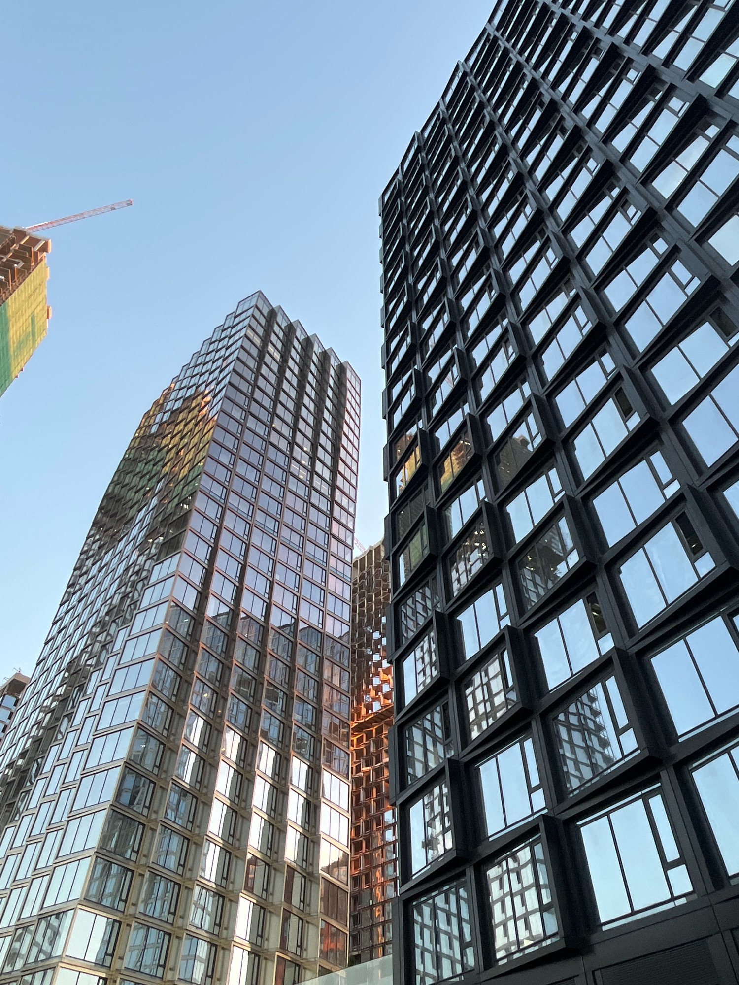 The image shows a low-angle view of modern skyscrapers against a clear blue sky. One building on the right has a dark facade with a grid of windows, and its reflective surface shows the sky and other buildings. To the left, another skyscraper with a glass facade, featuring a diamond-patterned design, reflects the sky and surrounding structures with warm tones. In the background, a construction crane is visible against the sky, attached to a building partially covered in green netting. The scene appears to be in an urban environment, likely a city under development. The lighting suggests it is daytime, possibly late afternoon, due to the warm reflections on the glass buildings. No people are visible in the image.