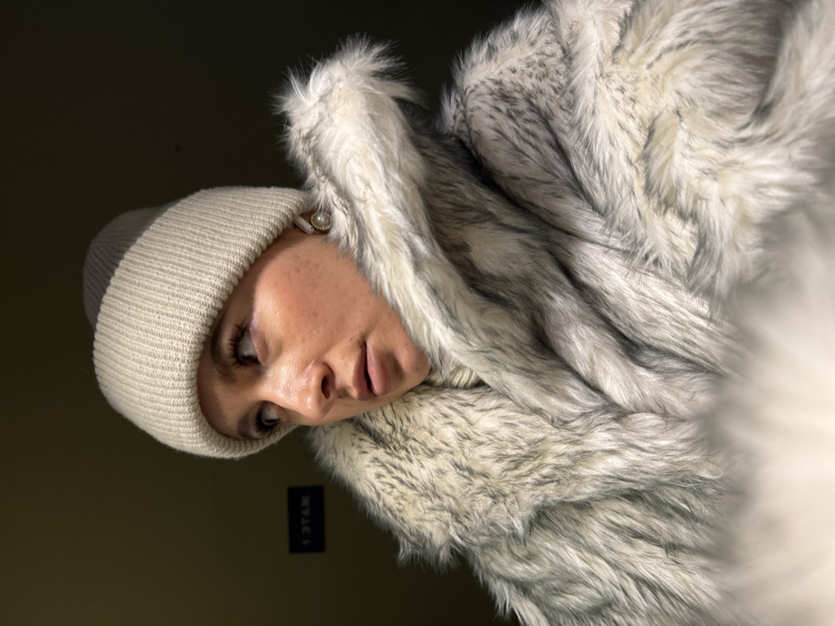 A female subject is depicted in a chest-up portrait within a dimly lit interior environment, located in Odintsovo, Russia. The subject wears a light beige ribbed knit beanie and a faux fur coat featuring shades of white, grey, and dark grey. A light-colored, ribbed scarf or turtleneck is visible beneath the fur collar. A single pearl earring with a gold-toned rim adorns her visible right ear. Her expression is neutral, with slightly parted lips, and her gaze is directed upwards and to the left. The background is dark and indistinct, featuring a dark rectangular sign on the left with light-colored, reversed text reading "Y TAЯM".