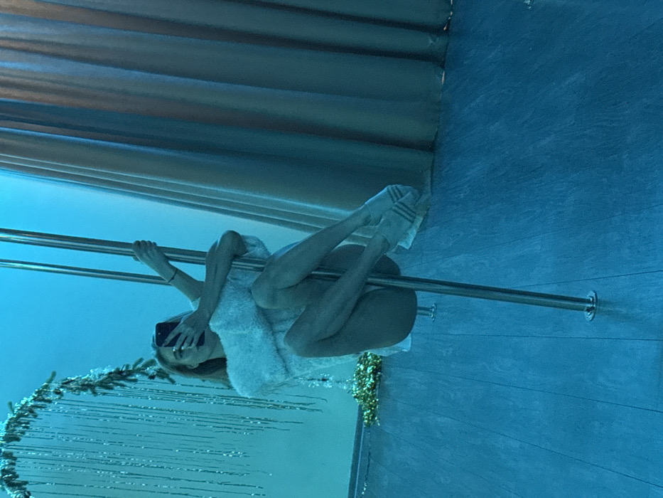 The image depicts a person performing on a metal dance pole within an indoor setting, illuminated by a pervasive blue light. The person is seen in a horizontal orientation within the frame, holding the pole with their hands and wrapping their legs around it in an acrobatic pose. They are dressed in light-colored, fuzzy or textured attire and wear white socks adorned with three dark stripes. In their left hand, the individual holds a black smartphone, aiming it towards themselves, indicating they are either taking a selfie or recording their performance. Their face is partially obscured by the phone.
The background features horizontal elements resembling blinds or panels across the top of the scene, and a textured wall on the right. In the lower left corner, a decorative garland or tinsel with shimmering strands is visible, suggesting a festive or ornamental environment. The dominant blue lighting casts a cool, almost aquatic or ethereal ambiance over the entire scene. There is no visible text in the image.