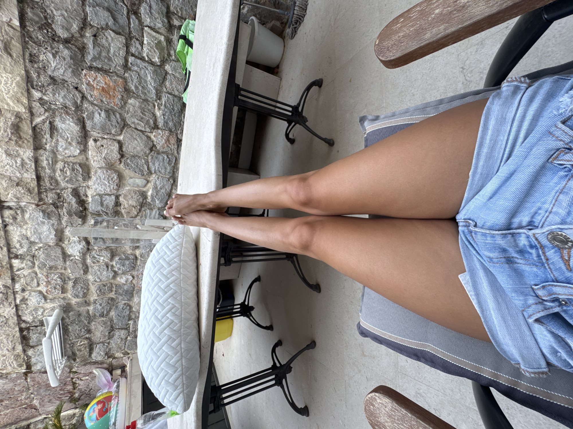 A person's legs are shown from the knees down, with their feet crossed at the ankles. They are sitting in a chair with wooden armrests and a striped cushion, wearing denim shorts. The legs are extended forward and rest on a surface that appears to be a table. The table has a white, textured top and is supported by two black, wrought-iron legs. Behind the table, a rough stone wall forms the background. Various objects are visible around the table and wall, including a white pillow on the table, a yellow bucket, a white bucket, a mop, and some inflatable toys on a counter. The ground is paved with light-colored tiles. The scene appears to be outdoors, possibly in a courtyard or patio area.