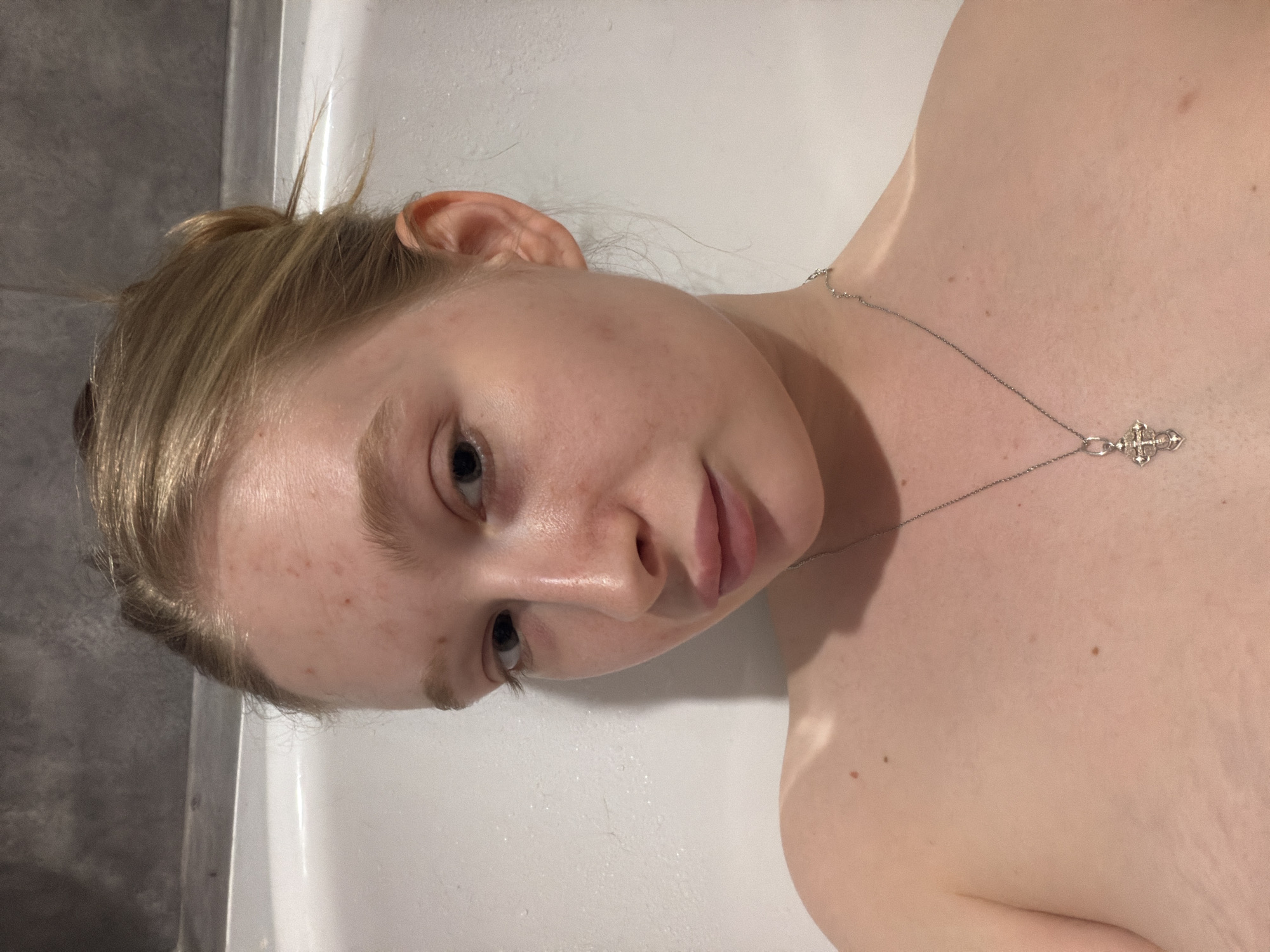 The image displays a young individual, likely female, with light blonde hair and fair skin, lying down in what appears to be a bathroom setting. The photograph is oriented on its side, showing the subject's head positioned on the left and their upper body extending towards the right. Their hair is partially tied back, with some wisps framing the face and crown.
The person has a neutral, relaxed expression, with their eyes looking towards the viewer. Visible on their face and upper chest are several small freckles or minor skin blemishes. A silver-colored chain necklace adorns their neck, featuring a prominent, intricately designed cross-like pendant resting on their chest.
The background consists of a smooth white surface, suggestive of a bathtub or shower wall, adjacent to a darker gray tiled wall on the far left. The lighting is consistent with an indoor, possibly artificially lit environment. Minor skin folds or stretch marks are noticeable on the shoulder and upper chest. There is no discernible text within the image. The depicted scene takes place indoors, within the context of Засечное, Russia.