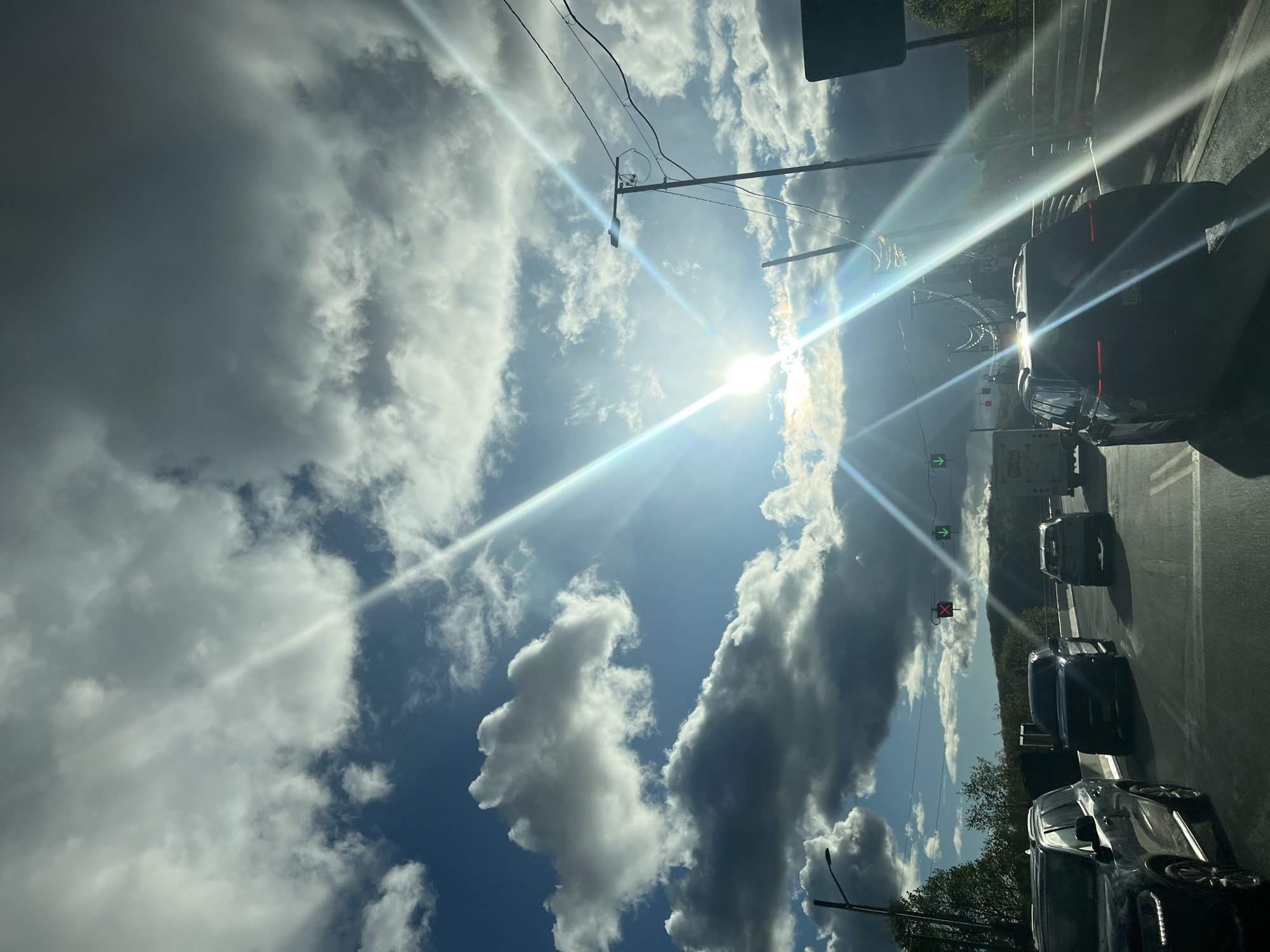 The image captures a bright, sun-drenched roadside scene, viewed from inside a vehicle looking forward. The sky dominates the upper two-thirds of the frame, showcasing a mix of fluffy white cumulus clouds and some darker, denser stratocumulus formations against a clear blue background. The sun is prominently featured, positioned high and slightly to the right of the center, emitting intense rays and significant lens flares that stretch across the image, highlighting atmospheric particles or glass surfaces.
Utility wires and poles are visible horizontally across the upper sky, typical of an urban or suburban setting. The bottom third of the image depicts a multi-lane road with several dark-colored vehicles, primarily SUVs and sedans, queued up in traffic. They appear stationary or moving slowly. To the right of the road, a green arrow traffic signal is visible, indicating a clear path forward, while a red "X" signal above it suggests a lane closure or restriction. Beyond the road, a line of trees and vegetation marks the horizon.
The overall impression is of a bright, clear day, likely mid-morning or afternoon, with strong sunlight creating dramatic glares. The scene suggests a busy road within the City of Poughkeepsie, United States of America. No specific people are visible, and while some license plates are present on the vehicles, any text on them is illegible due to distance and glare.