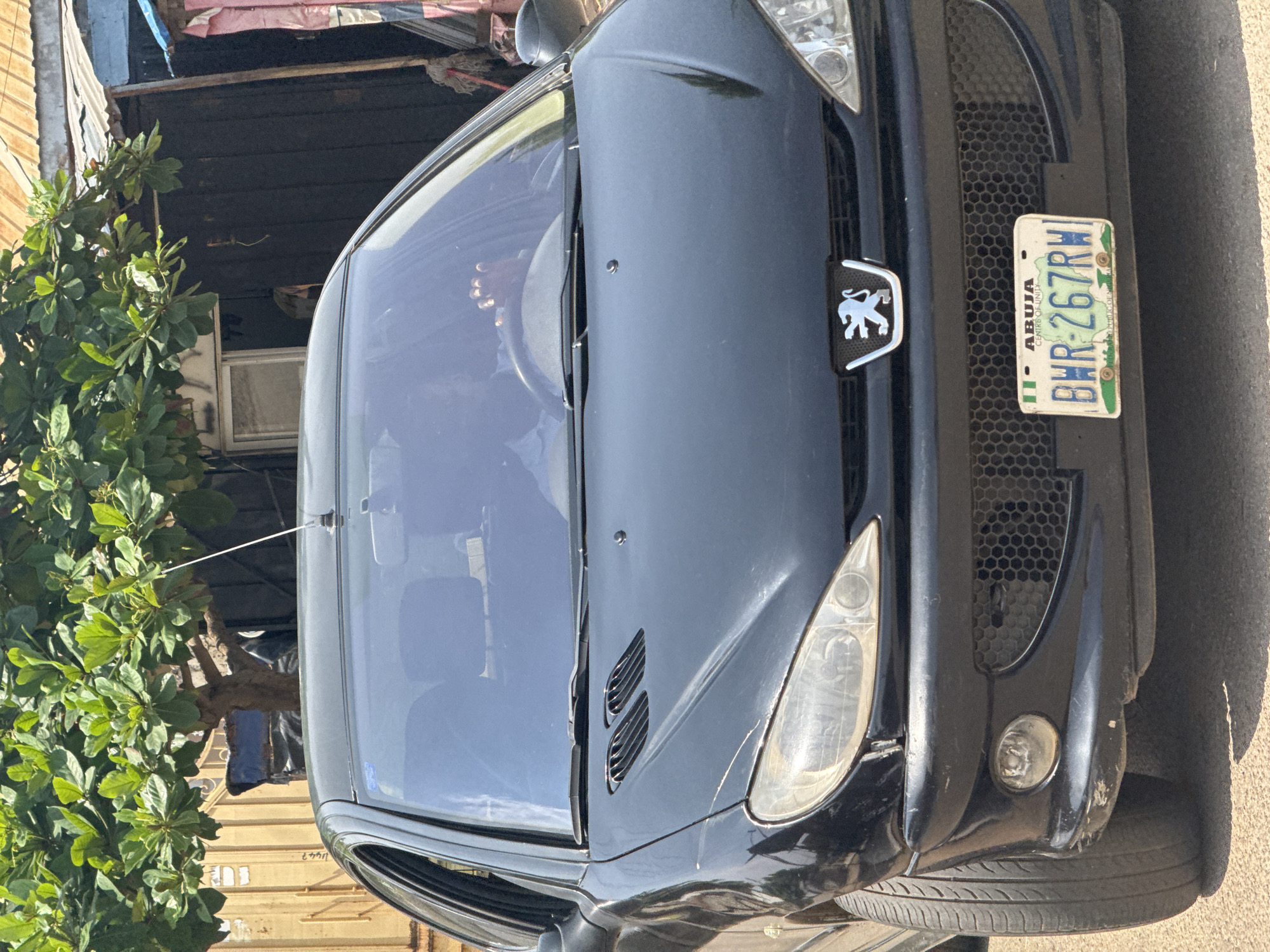A black Peugeot car is parked outdoors, facing the camera. The car is dark and reflects the sky. The front of the car features a license plate that reads "ABUJA BHR-267RW". To the left of the license plate is the Peugeot logo, a silver lion. The car's headlights and grill are visible. To the left of the car, a leafy green tree stands tall. In the background, there is a building with a corrugated metal roof and dark wooden walls. Sunlight suggests it is daytime. The scene appears to be in a residential or commercial area.