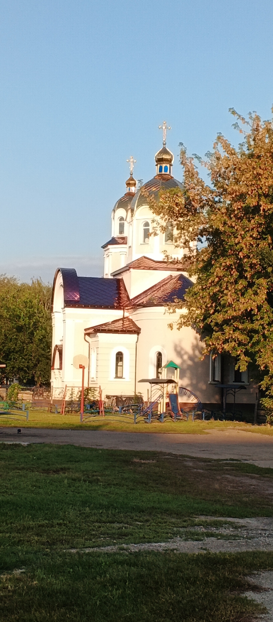 In Smila, Ukraine, a light-colored building with multiple domes and crosses, indicative of an Orthodox church, is visible. The building features arched windows and a dark, possibly copper, roof. In front of the church, a children's playground with blue and yellow structures, a slide, and a basketball hoop is present. The area is surrounded by green grass, trees with autumnal foliage, and a clear blue sky. The lighting suggests it is late afternoon or early evening.