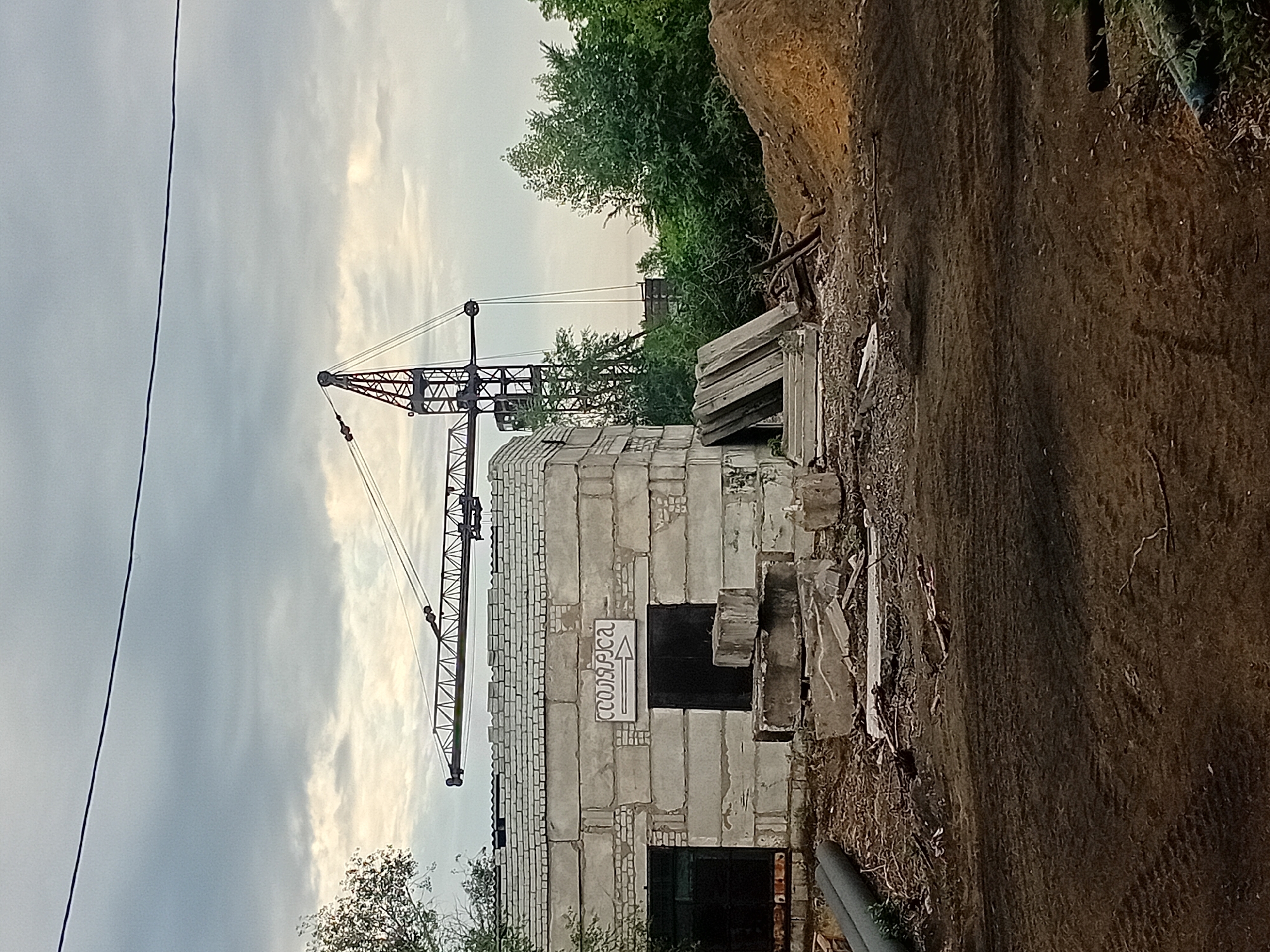 The image depicts a construction site in the city of Semey, Kazakhstan, under a cloudy sky. In the foreground, a dirt path with tire tracks suggests recent vehicle activity. To the left, a low-lying building constructed from concrete blocks is visible, with two dark entrances. Above these entrances, a sign with Cyrillic text and an arrow is mounted. Debris, including concrete slabs and pipes, are scattered around the building. In the background, a large tower crane is prominent, suggesting ongoing construction work. The scene is further characterized by dense green trees and a large mound of earth on the right side of the frame.