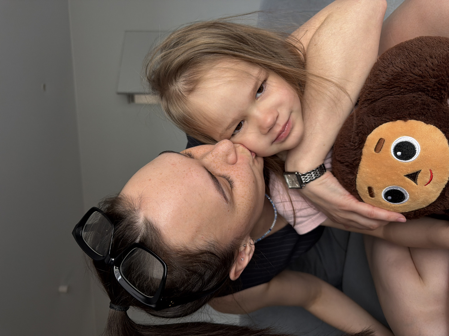 The image captures a warm, intimate moment between an adult female and a young child indoors. The adult, positioned with her head tilted, has dark hair tied back and wears black, square-framed glasses. Her eyes are closed as her face is pressed gently against the child's cheek, indicating a tender embrace or nuzzle. Freckles are visible on her skin. She is dressed in a dark top.
The child, with light brown hair, is seen embracing the adult, with her left arm wrapped around the adult's neck. She wears a pink top and a silver-colored watch on her left wrist. Her expression is soft and content, with a slight, gentle smile. In her right hand, she holds a brown, monkey-like stuffed animal with a light brown face, large black eyes, and a stitched red smile.
The background features plain, light grey walls, with a white, rectangular fixture (possibly a light or shelf) visible. A darker grey surface, likely a sofa or bed, is visible in the lower part of the frame. The overall atmosphere conveys affection, comfort, and a strong loving bond between the two individuals. The scene appears to be set in a home in Chaikovsky, Russia. There is no visible text in the image.