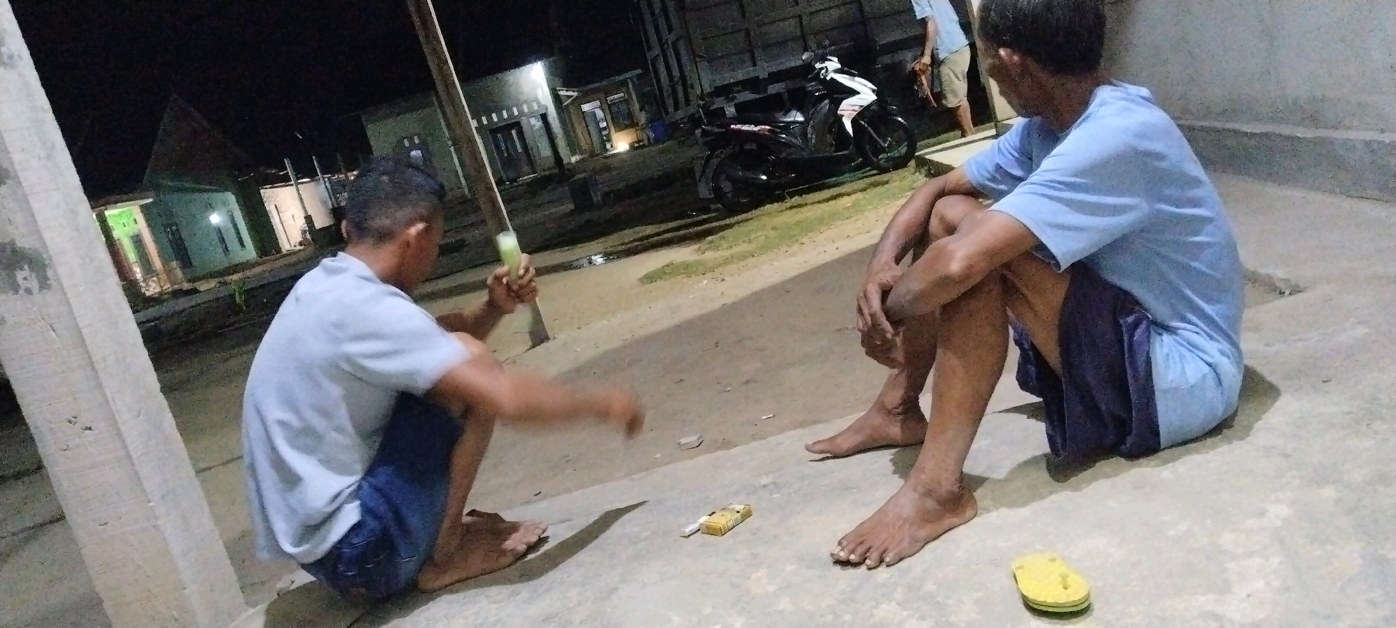 In Tanjung Ratu Ilir, Indonesia, two individuals are seated on a concrete ground at night. One person, on the left, is wearing a light blue t-shirt and dark shorts. This individual is crouched and holding a light green cylindrical object. In front of them, on the ground, is a pack of cigarettes and a single cigarette. The other person, on the right, is also seated, wearing a light blue t-shirt and dark blue shorts. They are facing away from the camera and resting their hands on their knees. A yellow flip-flop is visible on the ground near their feet. In the background, a black motorcycle is parked, and several buildings are illuminated. A wooden pole stands to the left of the person on the left. The overall scene suggests a casual, nighttime gathering.