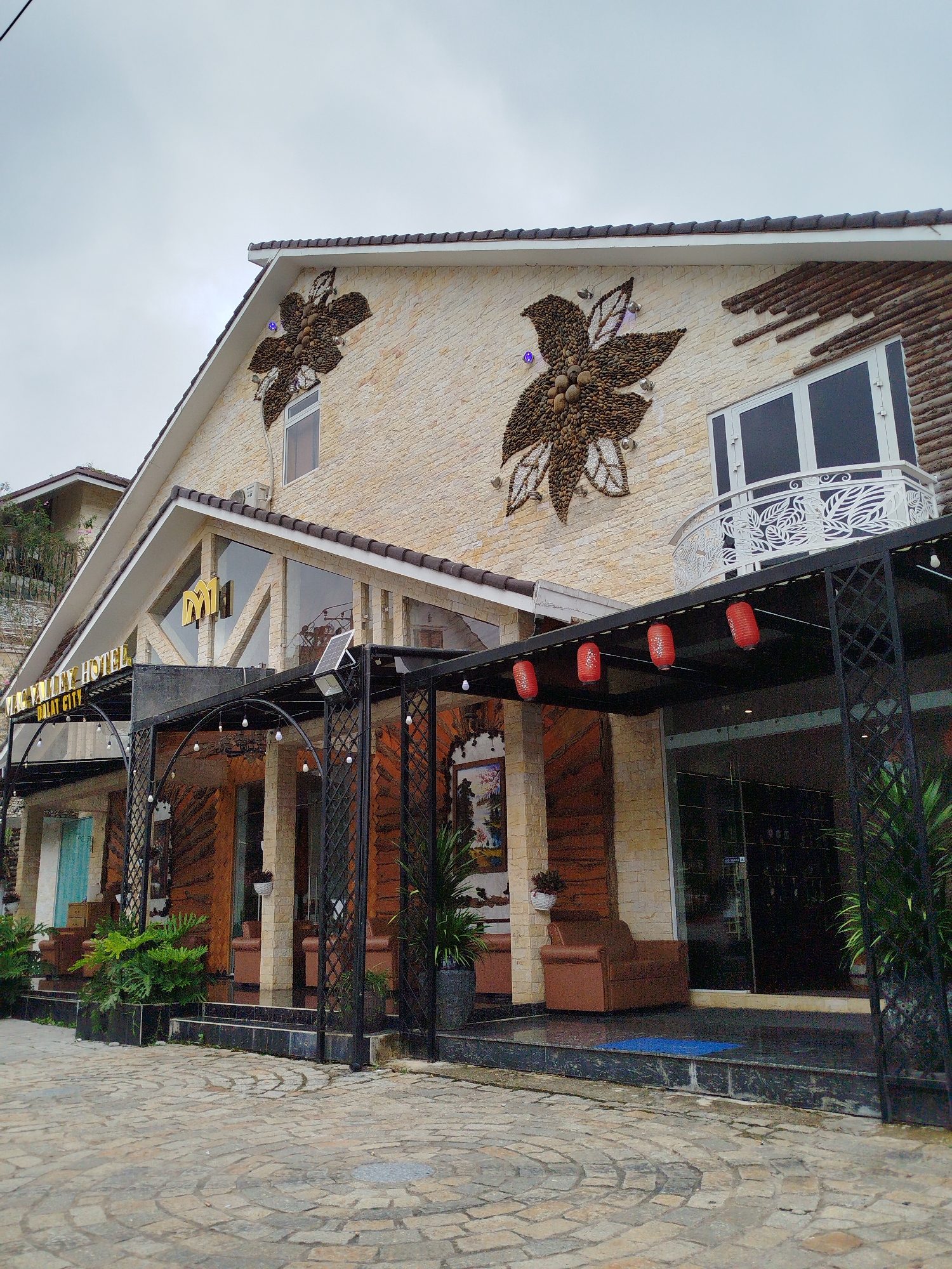The image depicts the exterior of the Mac Valley Hotel in Da Lat City, Vietnam. The hotel features a light-colored brick facade adorned with large, decorative floral motifs made from natural stones and pebbles. A prominent sign above the entrance reads "MAC VALLEY HOTEL" in golden lettering, with "DALAT CITY" below it. The entrance area is covered by a black metal canopy decorated with hanging string lights and three red lanterns. Several potted plants and outdoor seating areas with brown armchairs are visible in front of the hotel. The ground is paved with irregular light-colored stones. The sky is overcast.
