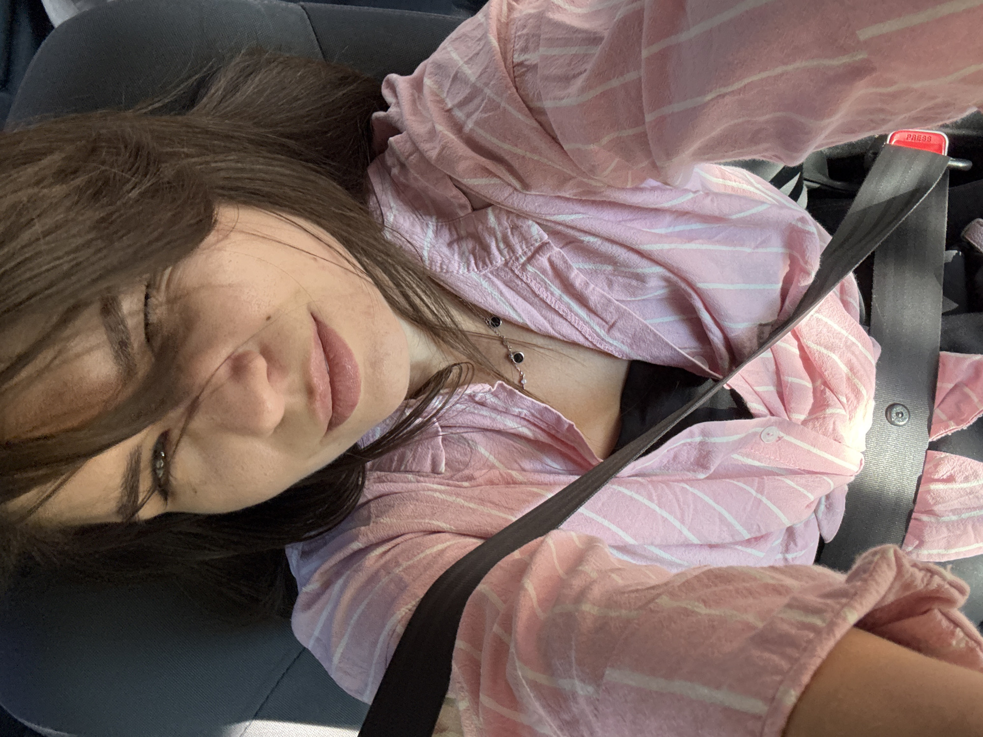 A woman with dark brown hair is seated in a car, wearing a pink and white striped shirt and a black garment underneath. A black seatbelt is fastened across her chest and torso. She is looking towards the camera with one eye partially closed in a wink. The background shows the interior of a car seat.