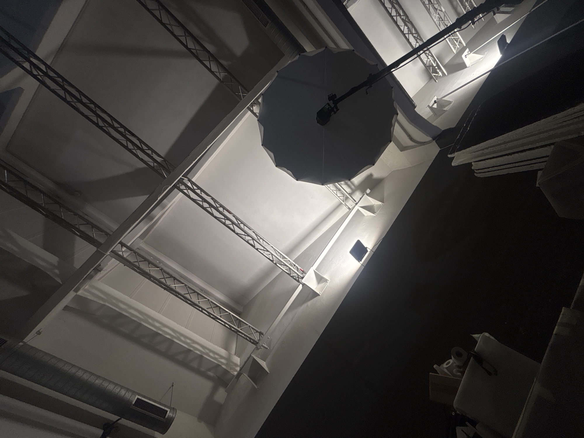The image shows a view looking up at the ceiling of what appears to be a large, industrial or commercial space. The ceiling is white, with exposed metal beams arranged in a grid pattern. An umbrella-shaped lighting apparatus is suspended from the ceiling. There's an air duct near the bottom-left corner. The right side of the image is obscured in darkness, with some shapes of objects visible. There are no people visible. The location in Milan, Italy is unknown.