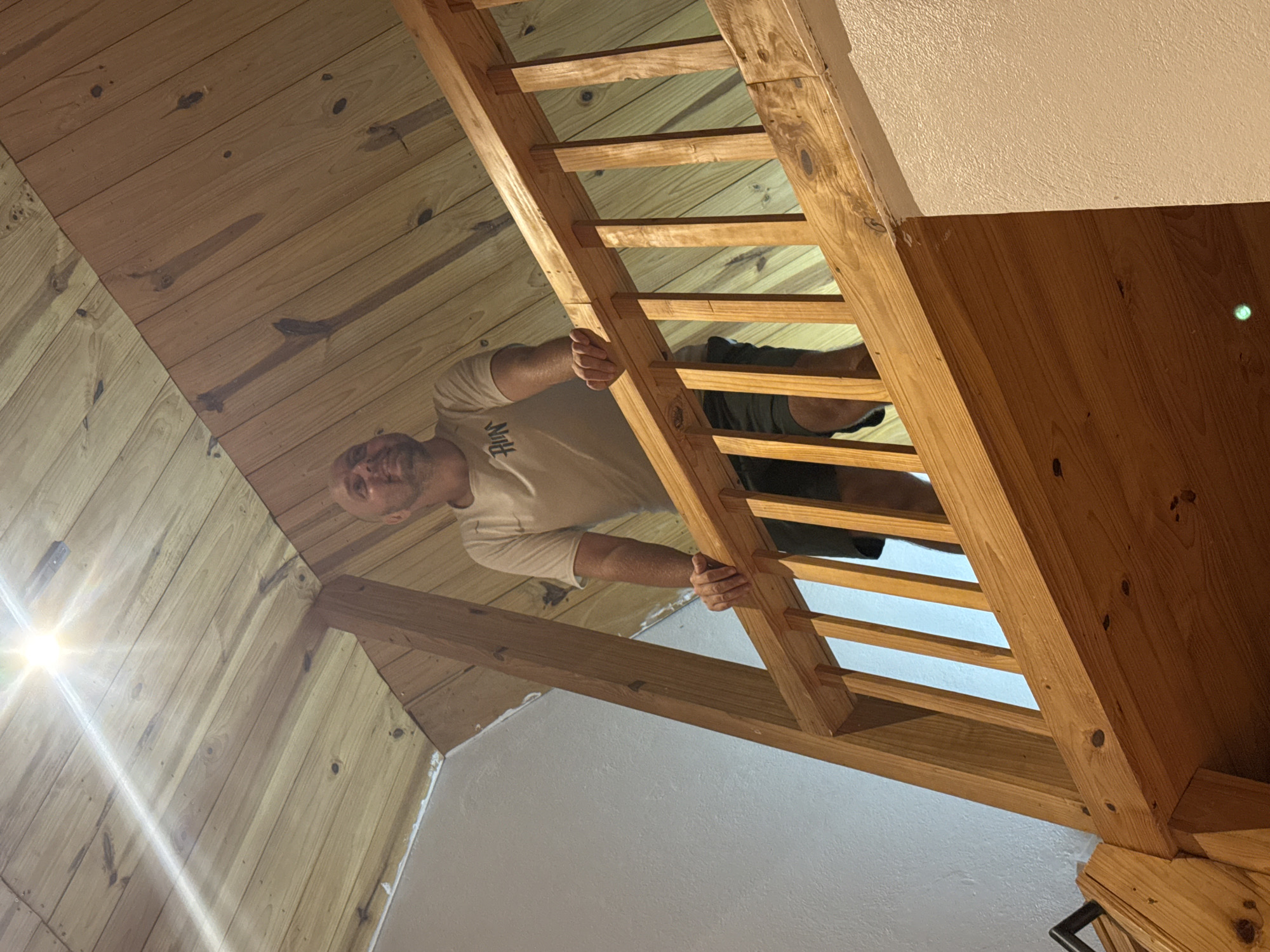 A man with short hair is standing on a wooden ladder inside a building. The building appears to have a wooden ceiling and white walls. The man is wearing a light-colored t-shirt and dark shorts. The environment appears to be indoors, with a rustic aesthetic. There is no information in the image to confirm the location is Du Già Commune, Vietnam.