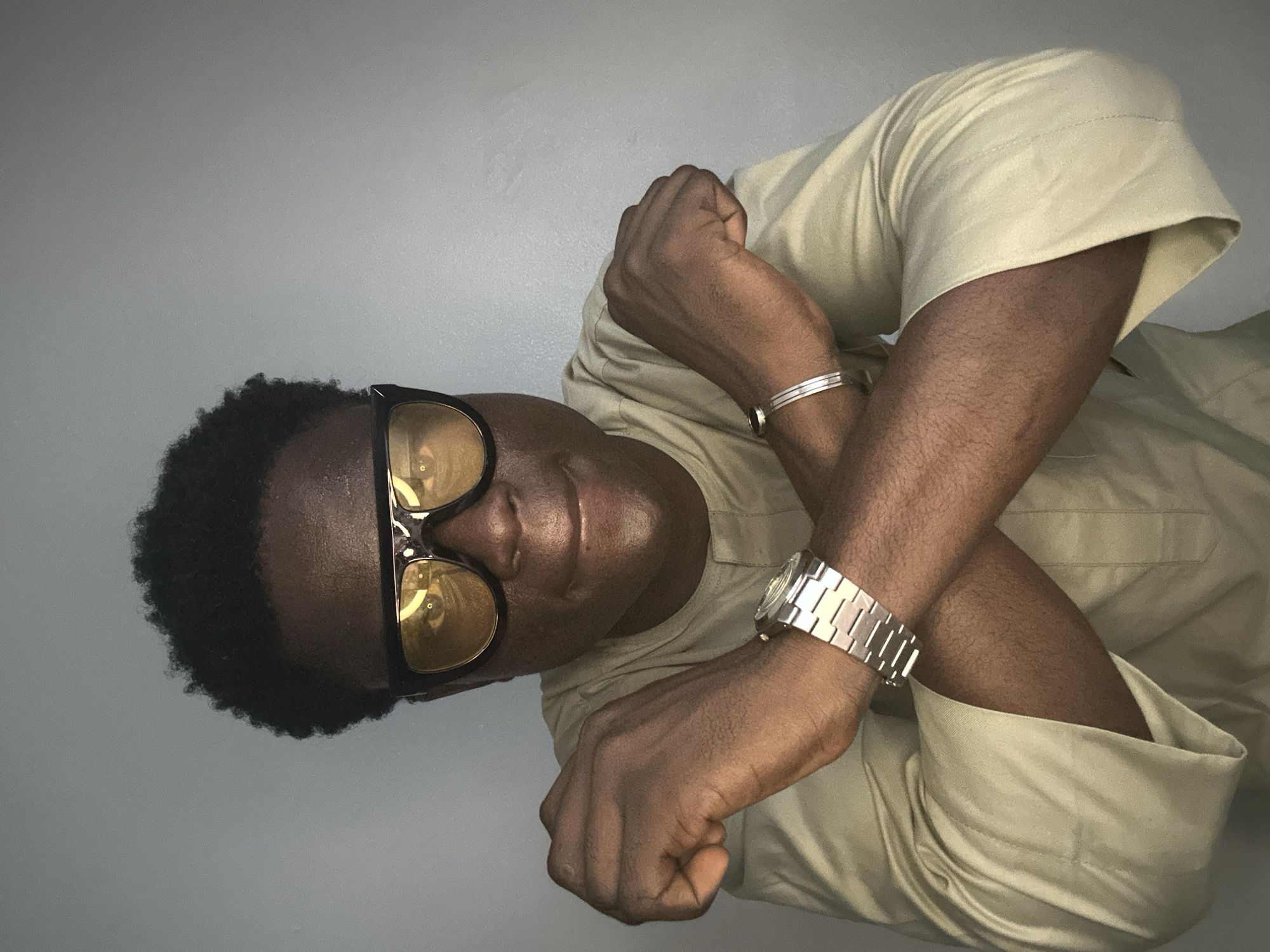 The media file features a portrait of a Black man posing against a plain, light-gray background. The man is wearing a light-tan, button-down shirt with subtle stitching details and a band at the collar, suggesting traditional Nigerian attire, potentially a buba. He has on sunglasses with thick black frames and yellow-tinted lenses. He is also accessorized with a silver watch on his left wrist and a thin silver bracelet on his right wrist. His arms are crossed in front of his chest. The lighting in the image is even, creating soft shadows that highlight the contours of his face and arms, and it appears to be indoors given the uniform lighting and backdrop. The overall mood is relaxed and confident, with the man looking directly at the camera with a slight smile. There is no visible text in the image. The style of clothing and the individual's features, combined with the provided location context of Badagry, Nigeria, suggest a contemporary portrait possibly taken in a domestic setting or a studio.