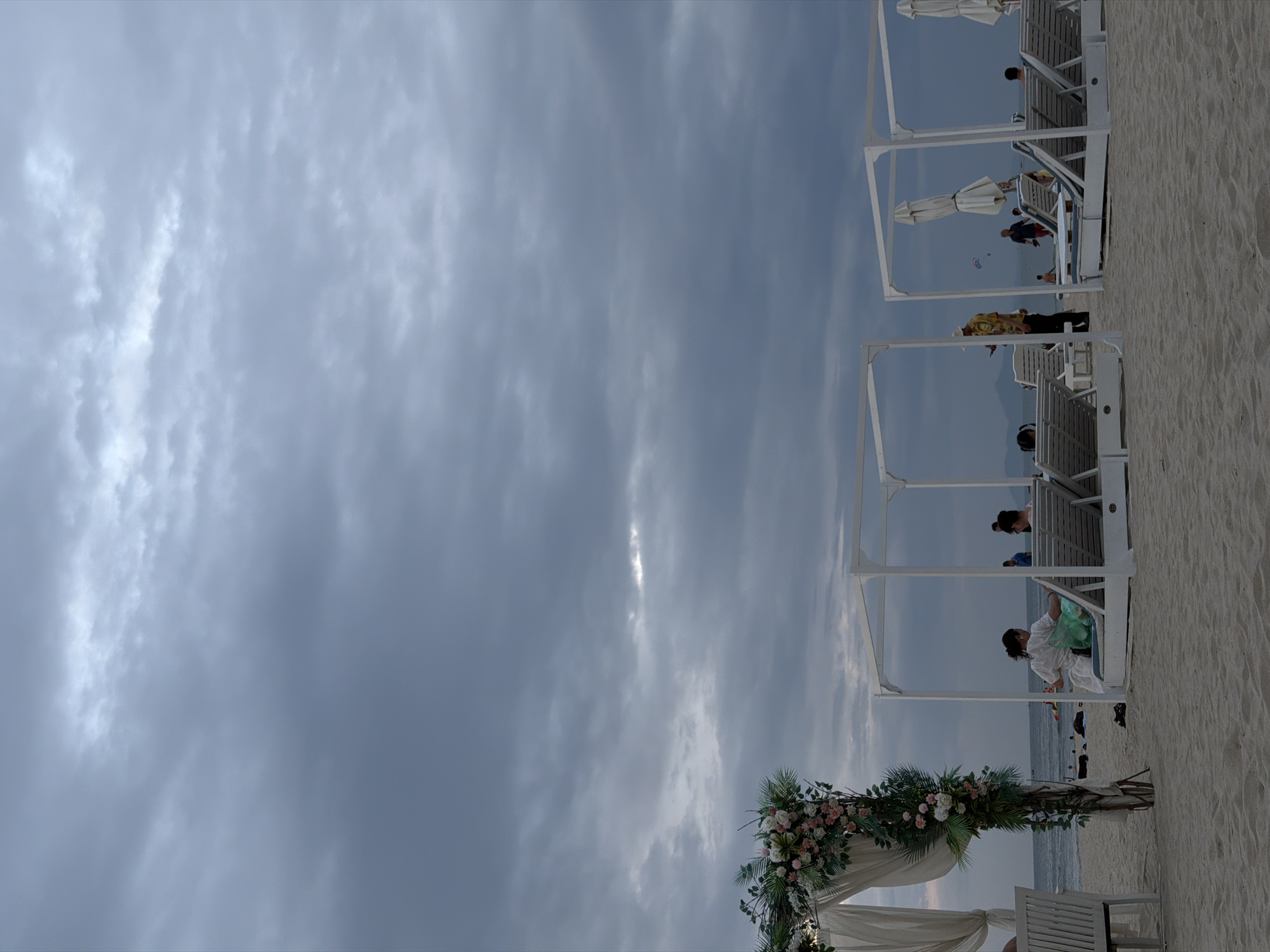 The image depicts a cloudy day at a beach, likely in Đà Nẵng, Vietnam, given the presence of typical beach amenities and the general atmosphere. In the foreground, there are several white cabanas with lounge chairs, some occupied by people. To the left, a decorated archway with flowers suggests a special event, possibly a wedding. The beach itself is sandy, and the ocean is visible in the background, with some small boats and distant landforms. The sky is dominated by overcast clouds, casting a subdued light on the scene. A few individuals are seen walking on the beach and in the water. One person in a colorful shirt stands near a cabana, looking at their phone. Another person is seen parasailing in the distance. The overall impression is a relaxed, possibly end-of-day setting at a tourist beach.